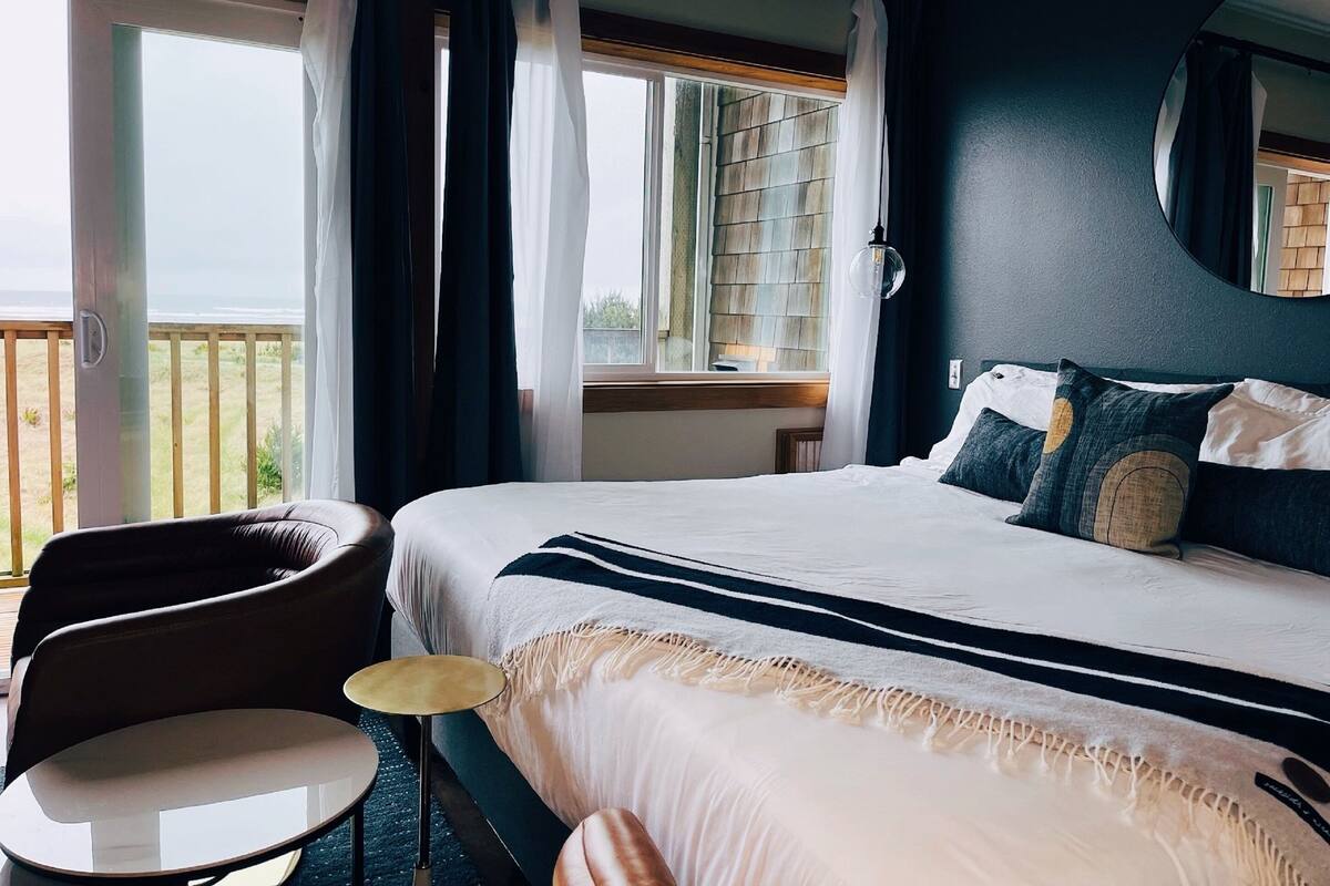 Quarto com cama grande, uma cadeira, uma mesa redonda e vista para o exterior.