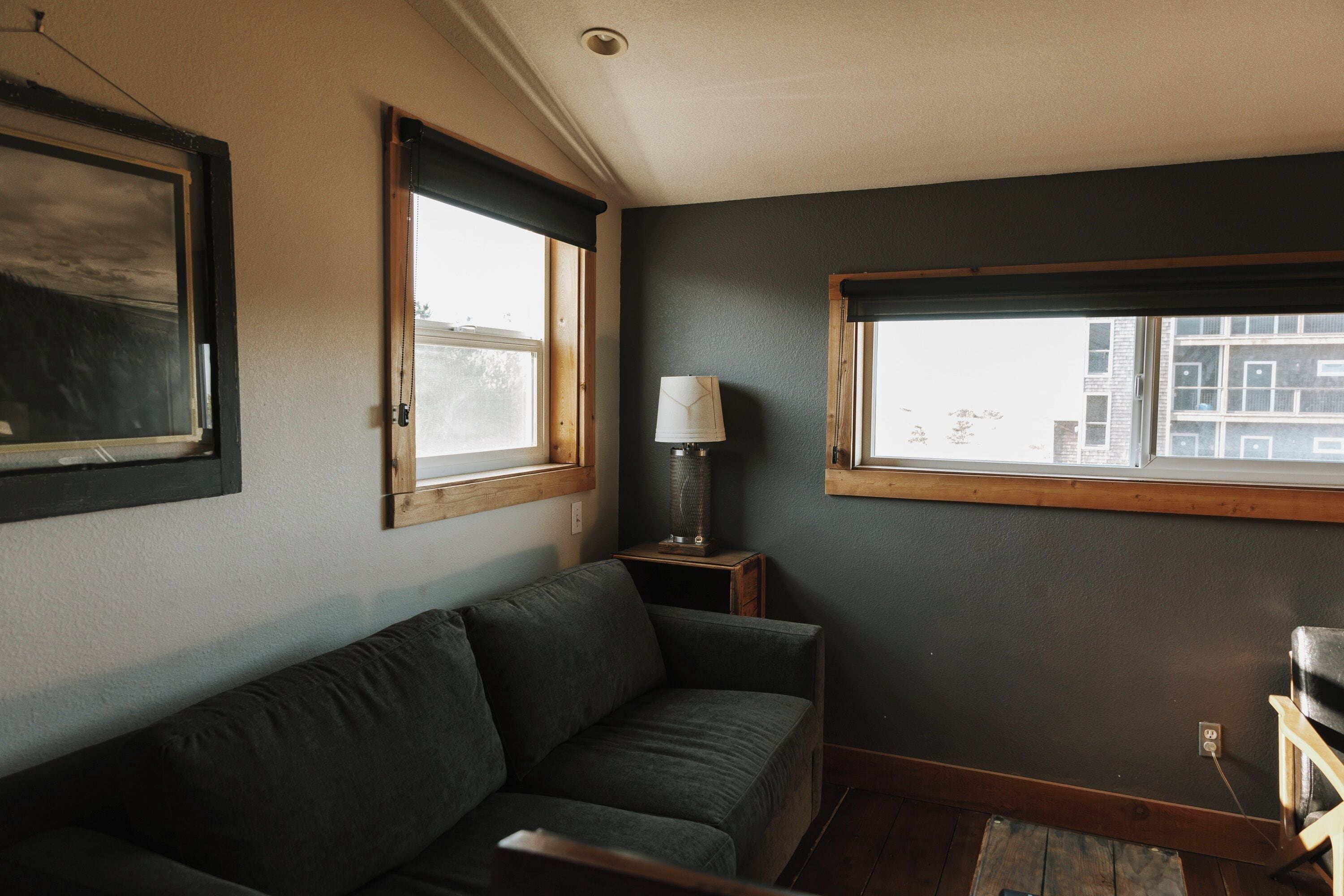 A room with a dark-colored sofa, a lamp on a small table, and a window with a view of buildings.