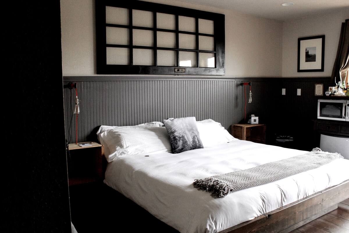 Quarto bem arrumado com uma cama grande, mesa de cabeceira, prateleira lateral, televisão fixada na parede e janela com persianas.