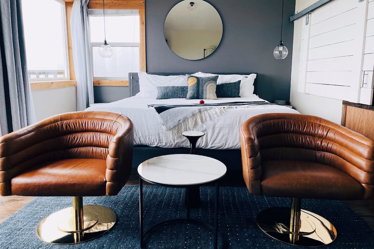 Quarto de hotel com uma cama, duas poltronas de couro, uma mesa pequena redonda, um espelho redondo e uma janela com cortinas.