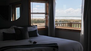 Un dormitorio con un ventanal que da a una playa, una cama con almohadas y una mesita de noche.