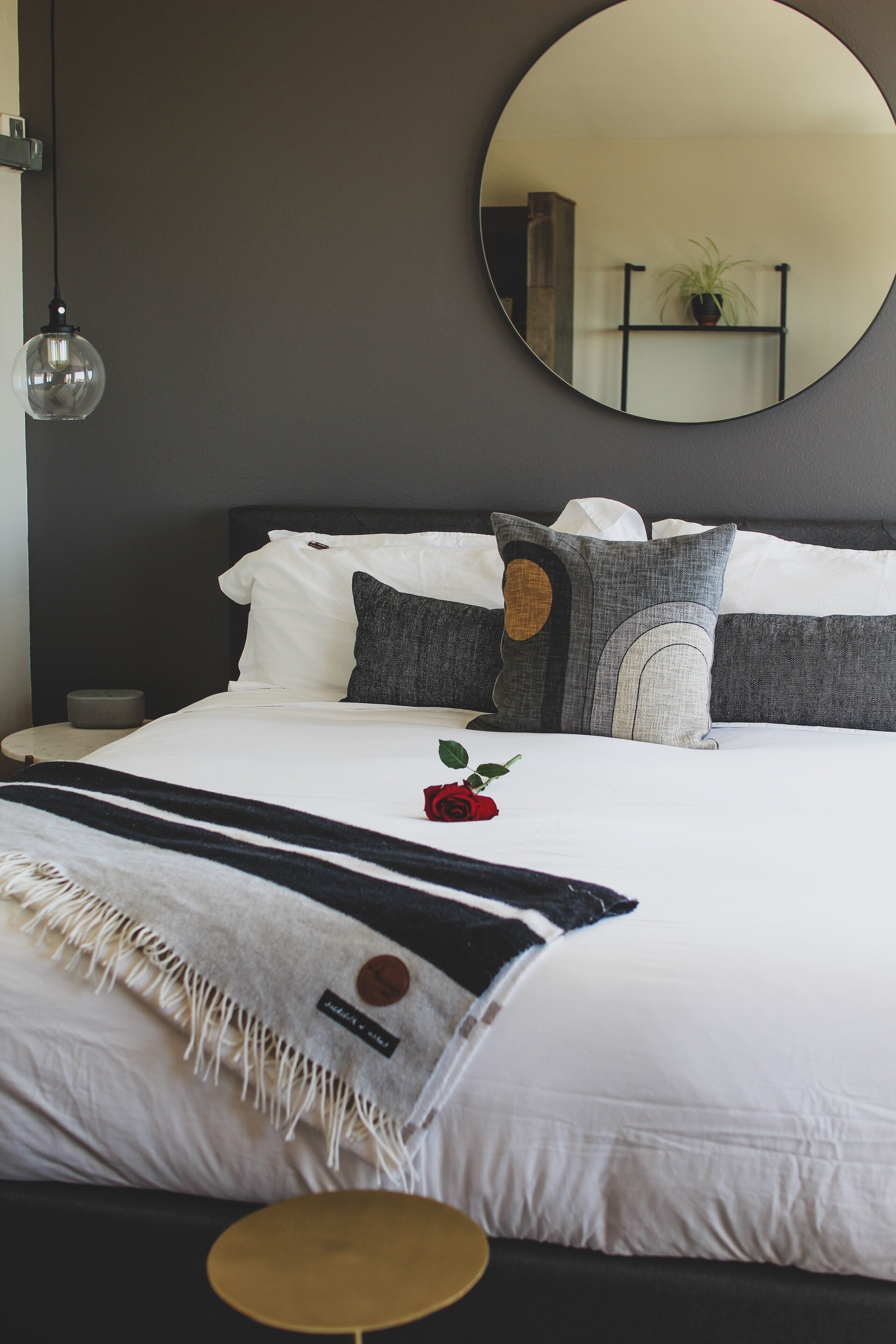 A neatly made bed with white and gray bedding, a round mirror above it, and a small round table beside the bed.