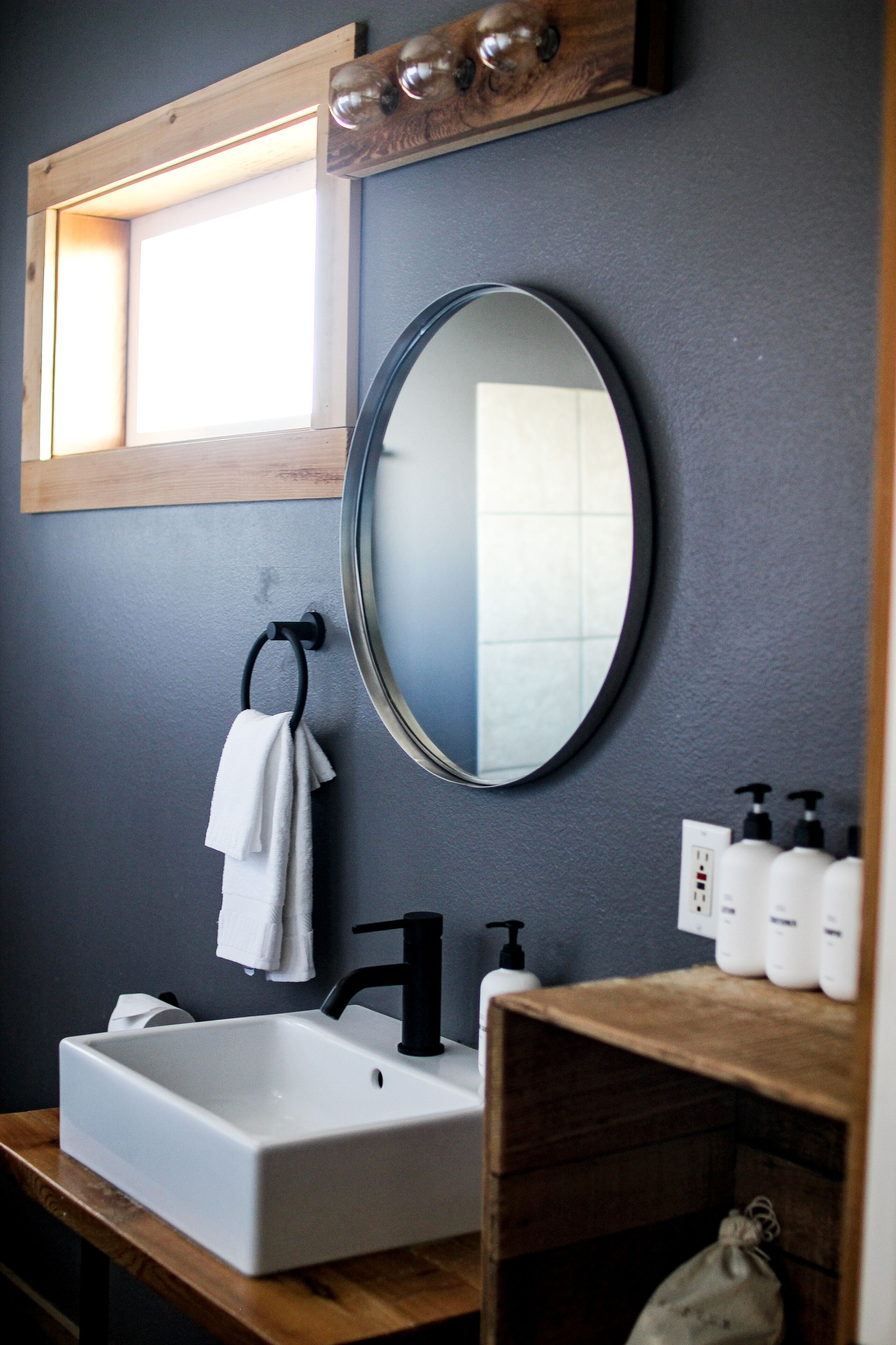 A bathroom with a white sink, black faucet, and a round mirror. There is a wooden shelf with toiletries and a towel hanging on a hook.