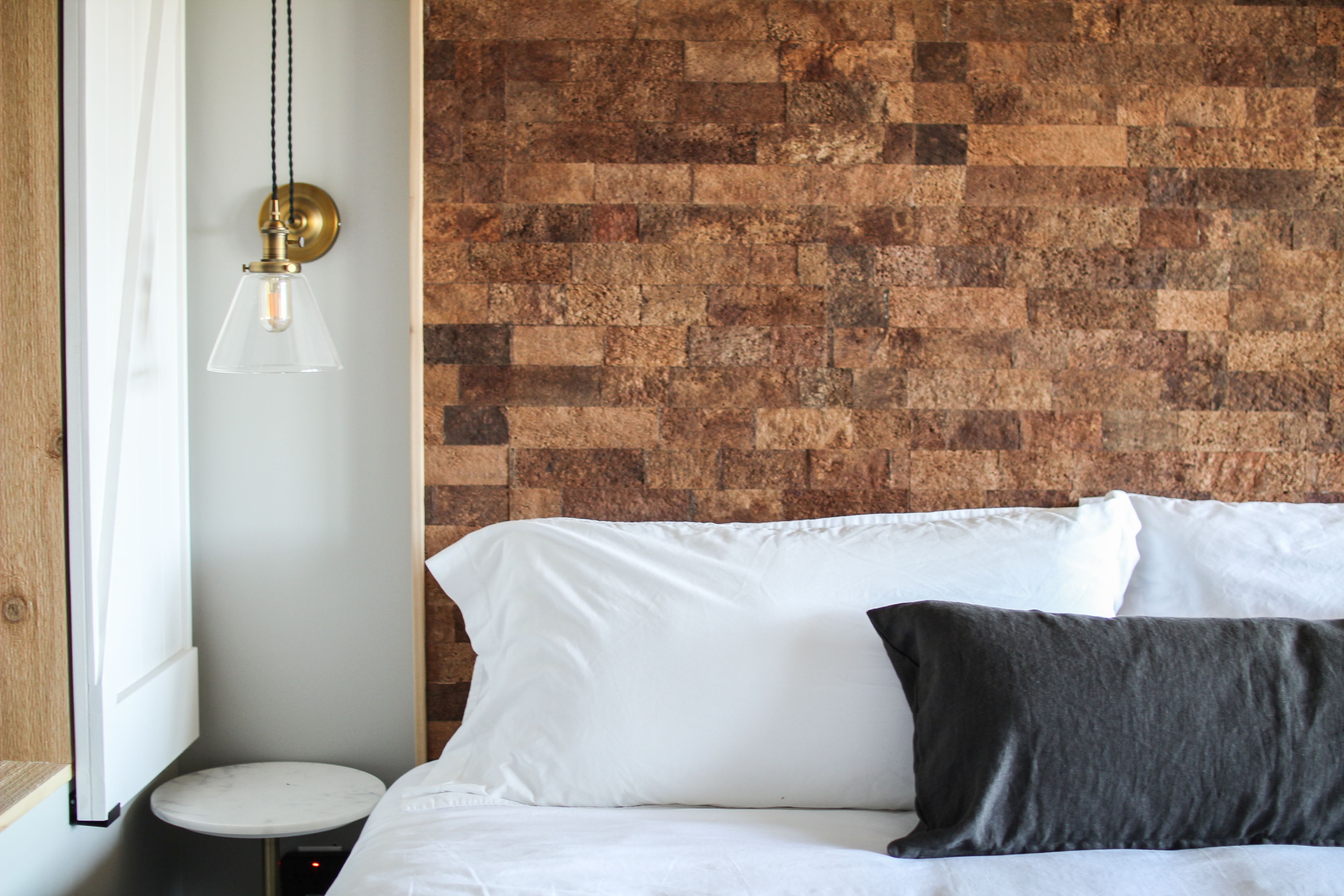 A bedroom with a brick-patterned wall, a bed with white and dark pillows, a round table, and a hanging light fixture.