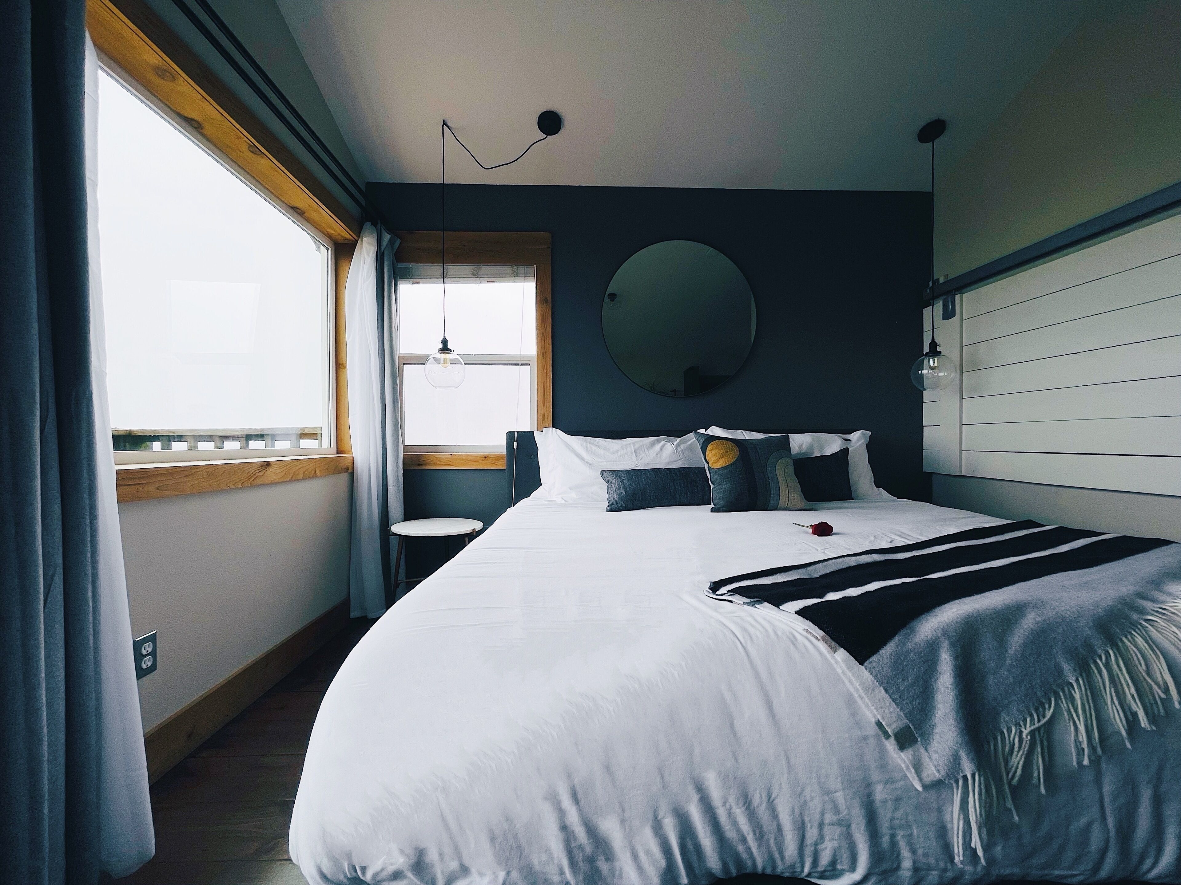 A bedroom with a large bed, a round mirror, a small stool, and a window with curtains.