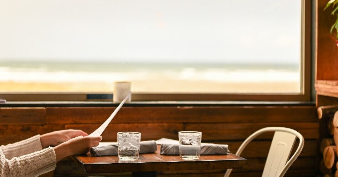 Eine Person hält eine Speisekarte an einem Holztisch, wo zwei Gläser Wasser und eine Tasse stehen. Im Hintergrund ist durch ein Fenster der Strand zu sehen.