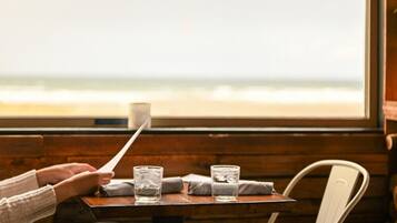 Eine Person hält eine Speisekarte an einem Holztisch, wo zwei Gläser Wasser und eine Tasse stehen. Im Hintergrund ist durch ein Fenster der Strand zu sehen.