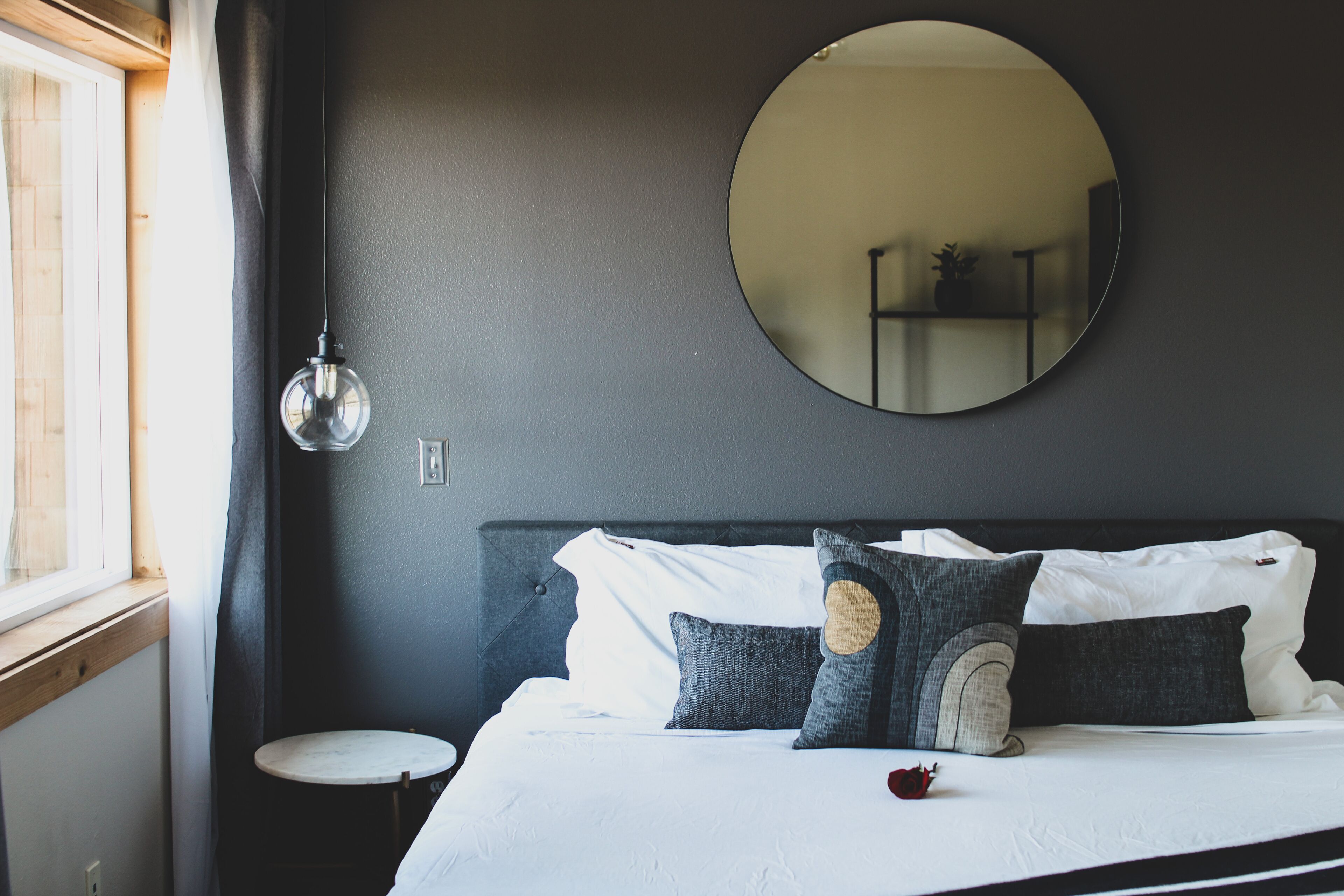 Una cama bien hecha con sábanas blancas y varios almohadones, un espejo redondo en la pared y una mesita de noche.