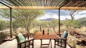 An outdoor wooden table with two chairs, a bottle of wine, and a glass, set against a backdrop of a natural landscape with trees and mountains. - Waterberg Guest Farm (Otjiwarongo)
