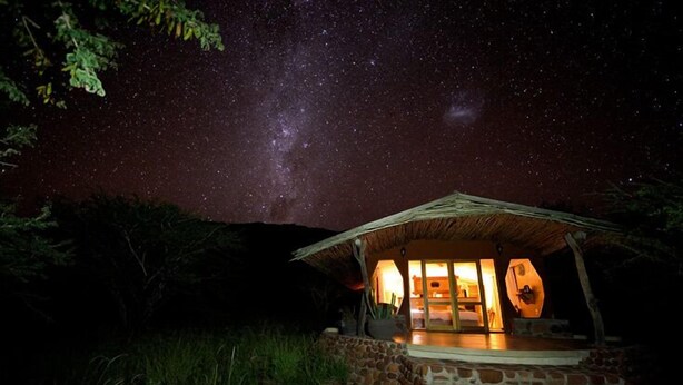 A thatched-roof structure with a stone foundation, a glass door, and a view of the night sky with stars and the Milky Way. - Waterberg Guest Farm (Otjiwarongo)