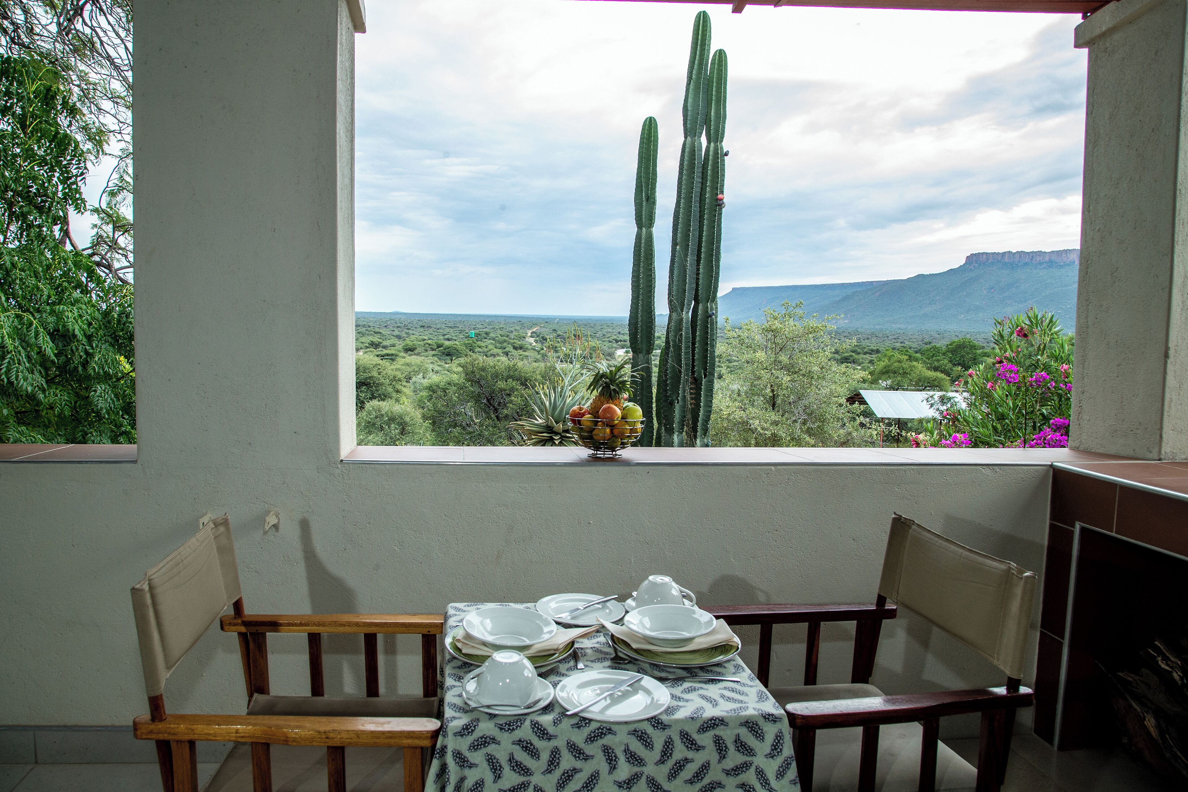 A dining area with a table set for four, a window with a view of cacti and a mountain, and a plant with purple flowers on the windowsill.
