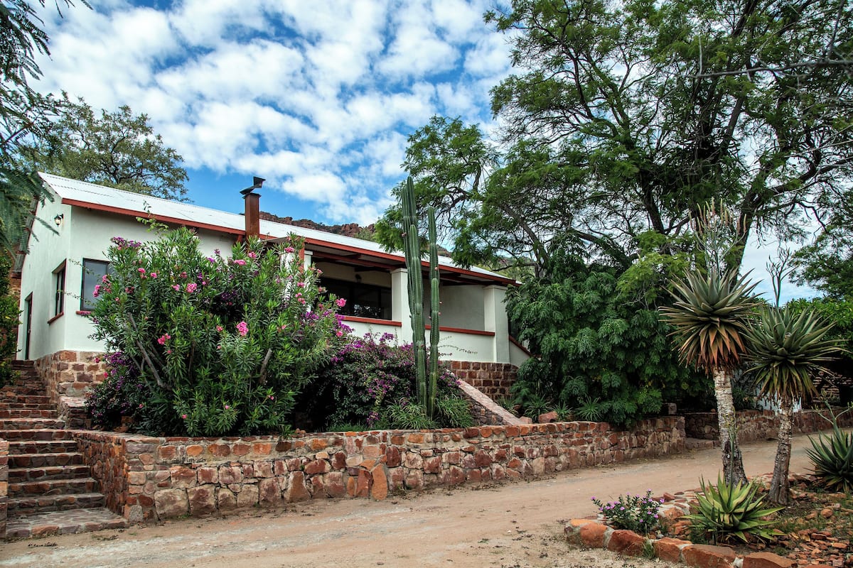 A two-story house with a stone staircase leading to the entrance, surrounded by various plants and trees.