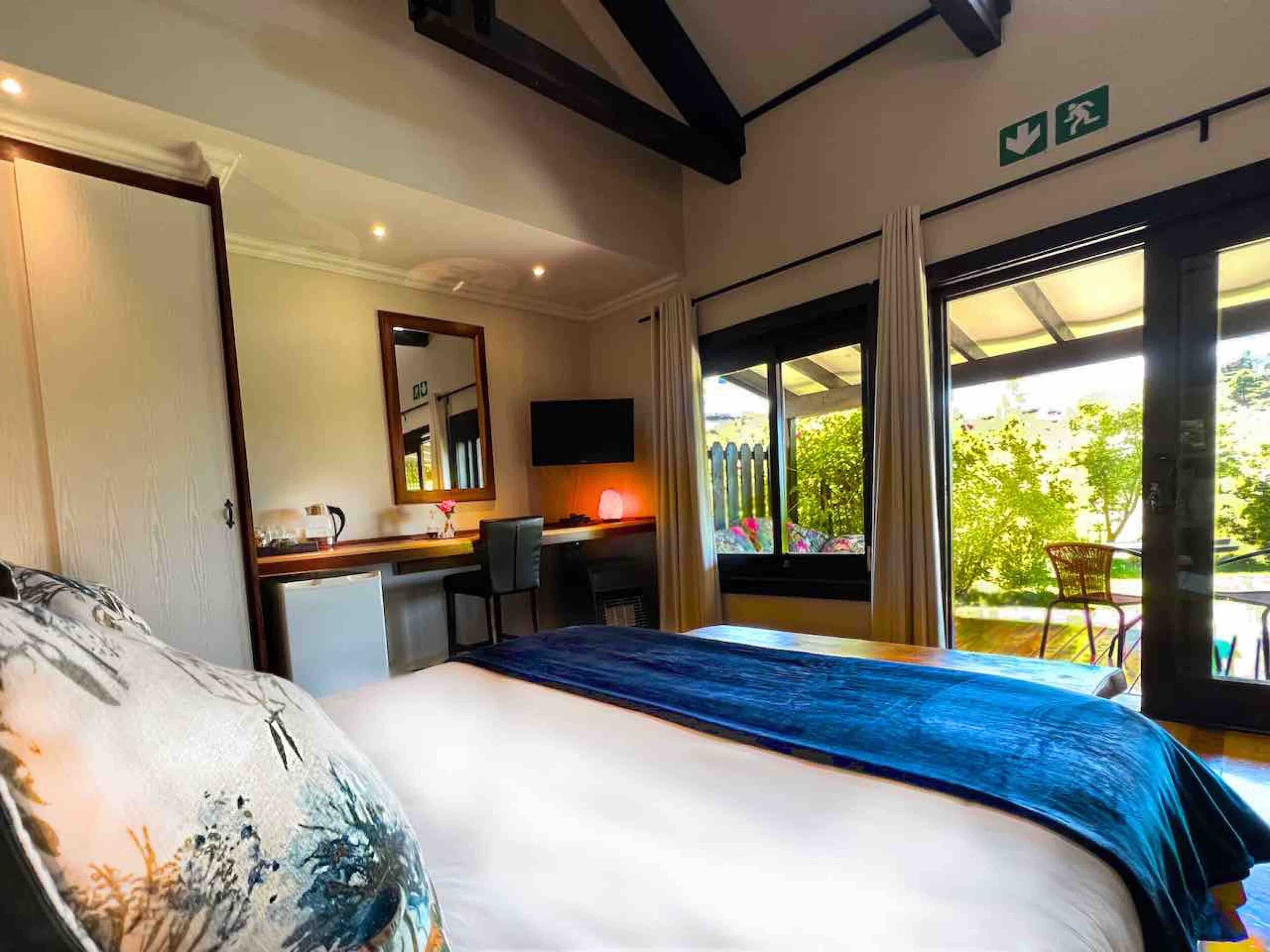 A hotel room with a bed, a desk, a chair, a TV, and a balcony with a view of trees.