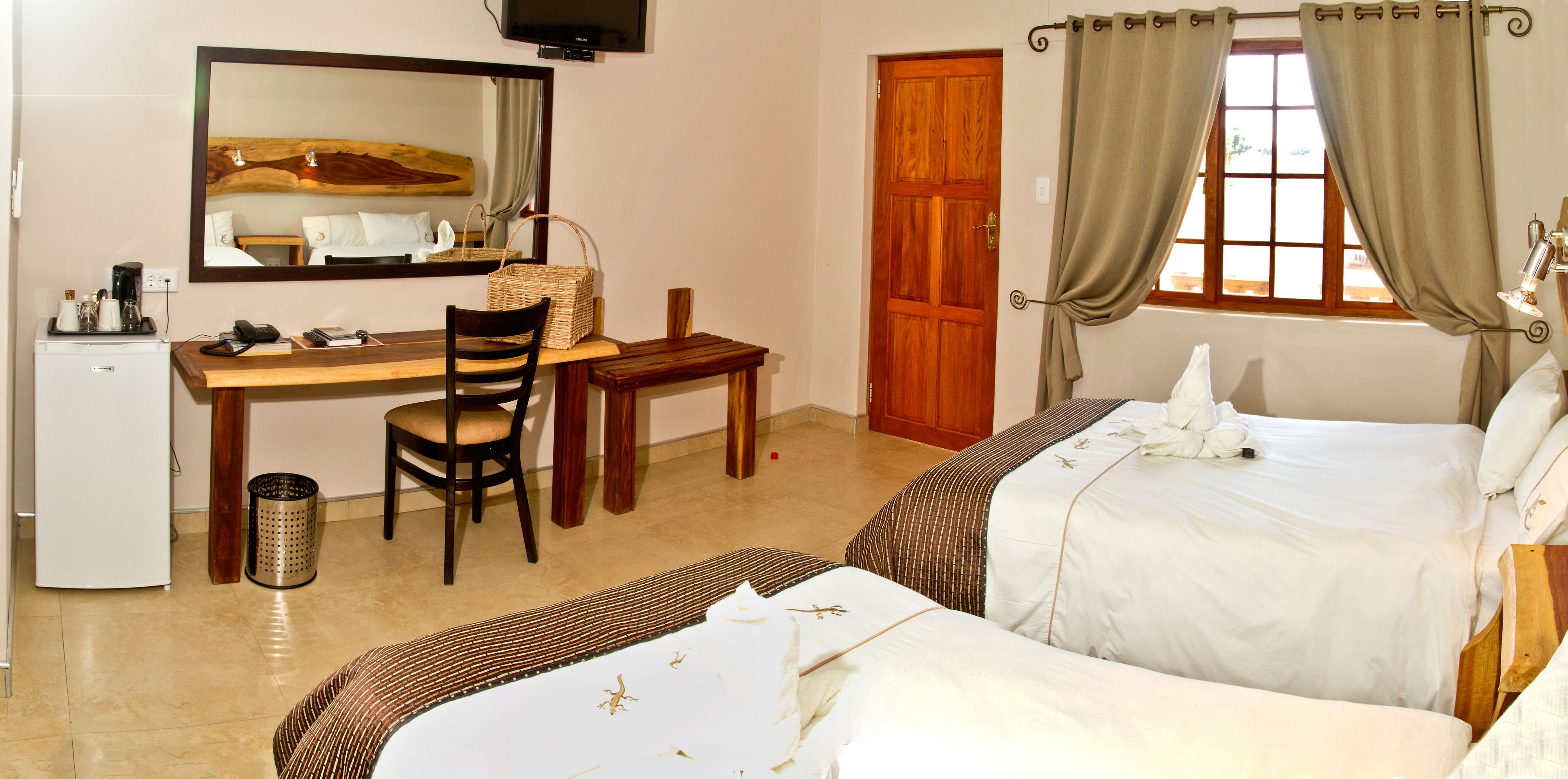 Habitación de hotel con cama, escritorio y silla, refrigerador pequeño, espejo y ventana con cortinas.