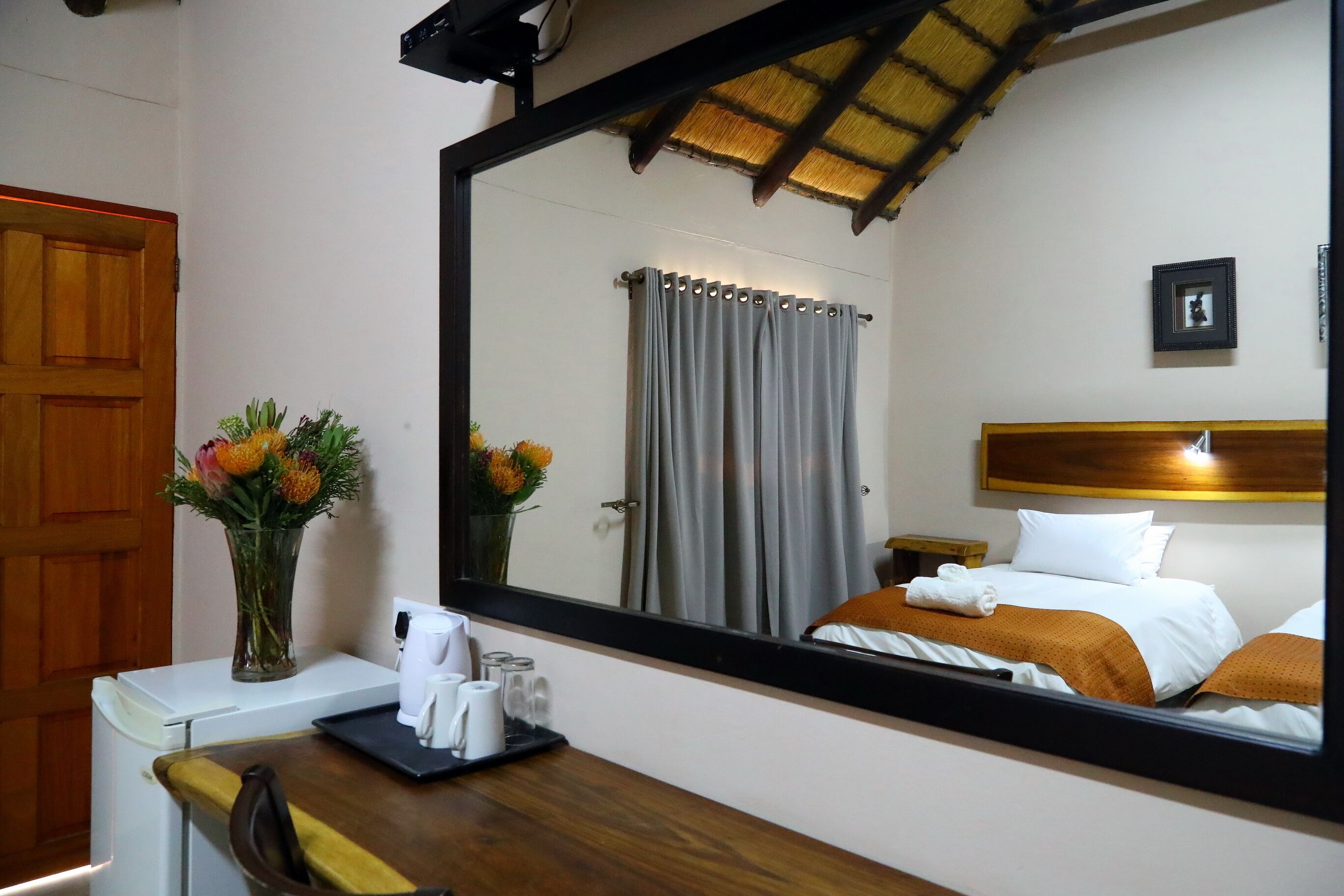 Habitación de hotel con un espejo grande que refleja una cama con ropa de cama blanca y naranja, un cabecero de madera y una mesita de noche con una lámpara. Se ve un escritorio de madera con una silla, un florero y artículos de tocador.