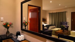 Une chambre d’hôtel avec un grand miroir, une table basse en bois, un canapé, un fauteuil et un lit agrémenté d’un rideau vert.