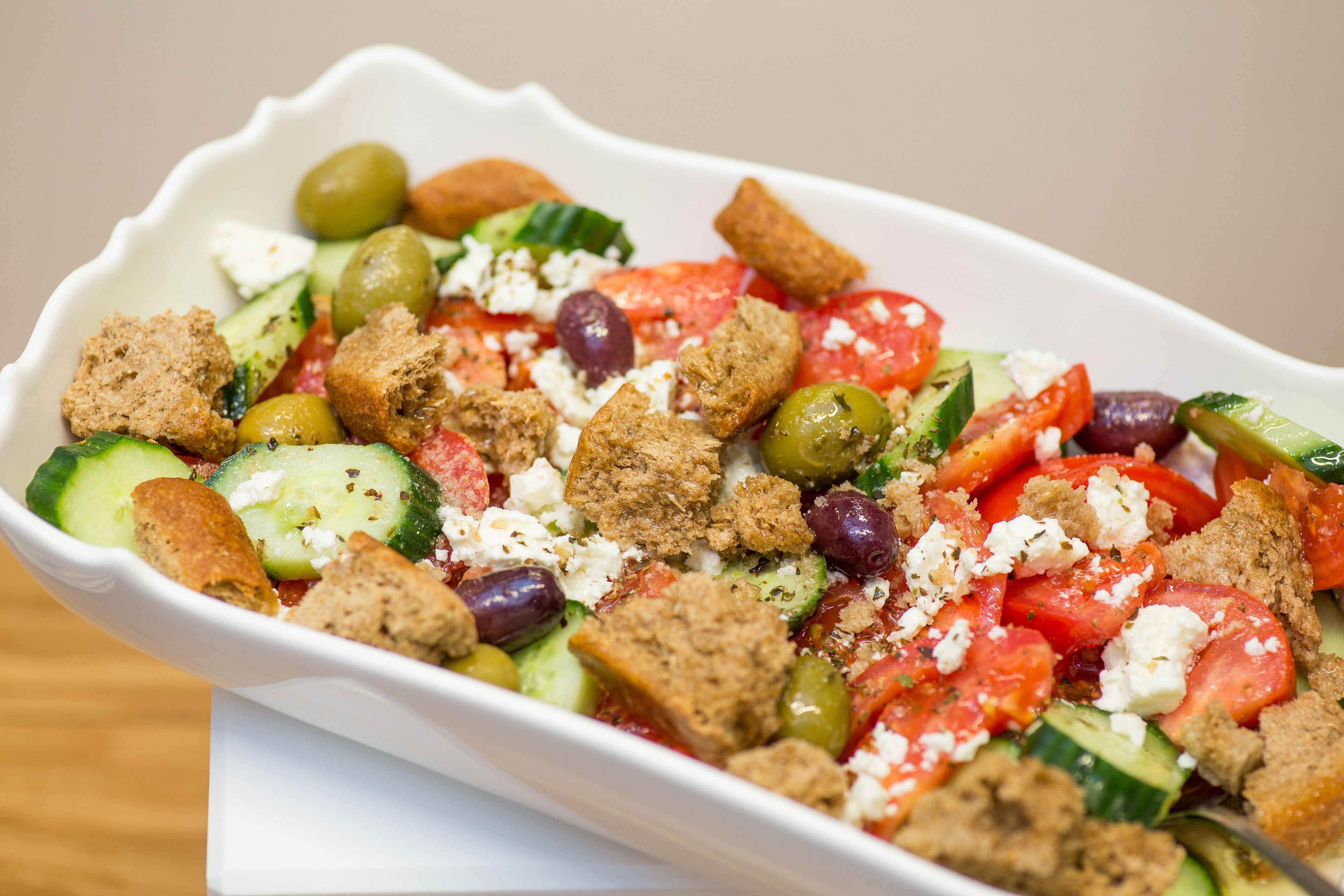 Un recipiente blanco rectangular con una ensalada de pepinos, tomates, aceitunas, queso feta y crotones.