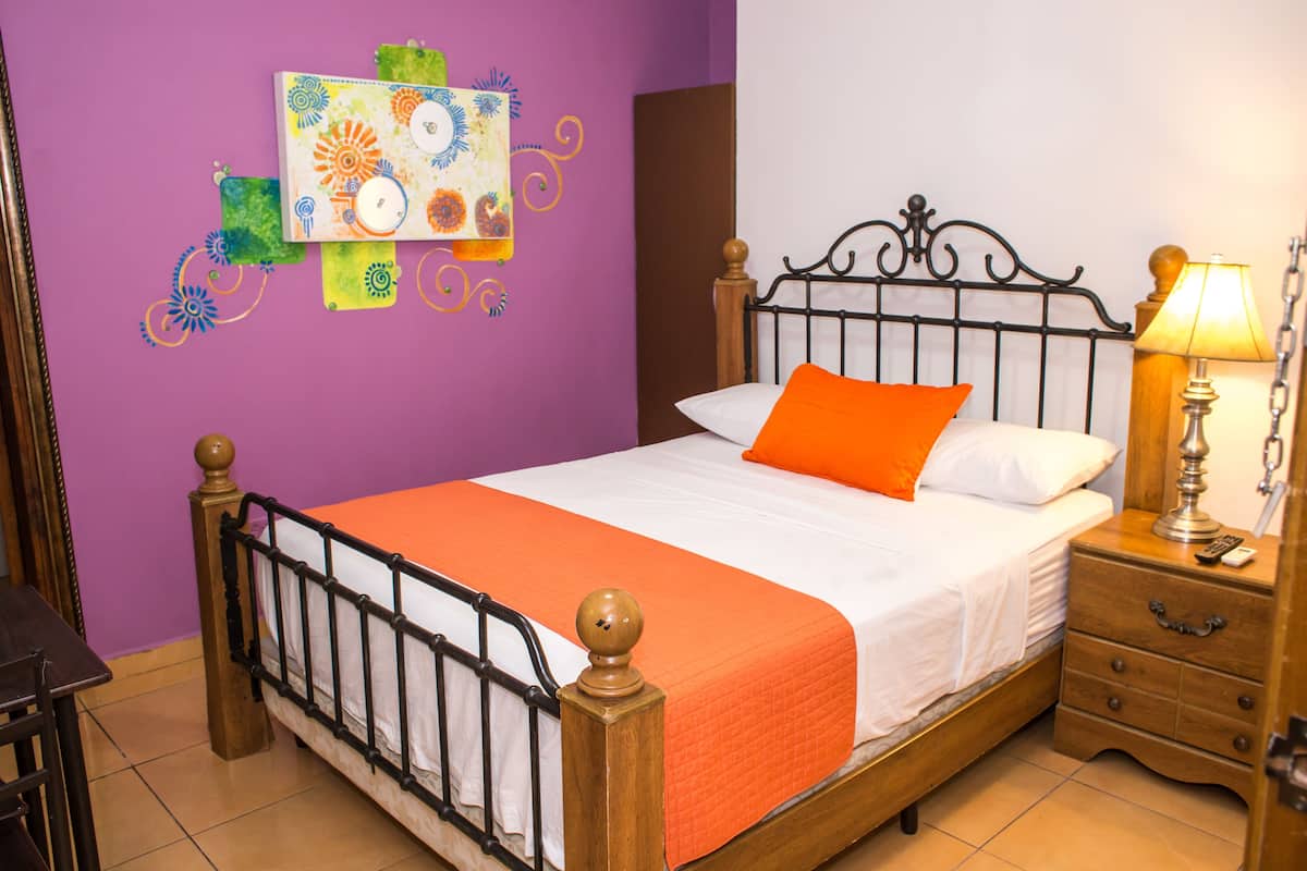 A neatly made bed with an orange blanket and pillows, a wooden nightstand with a lamp, and a wall decoration.
