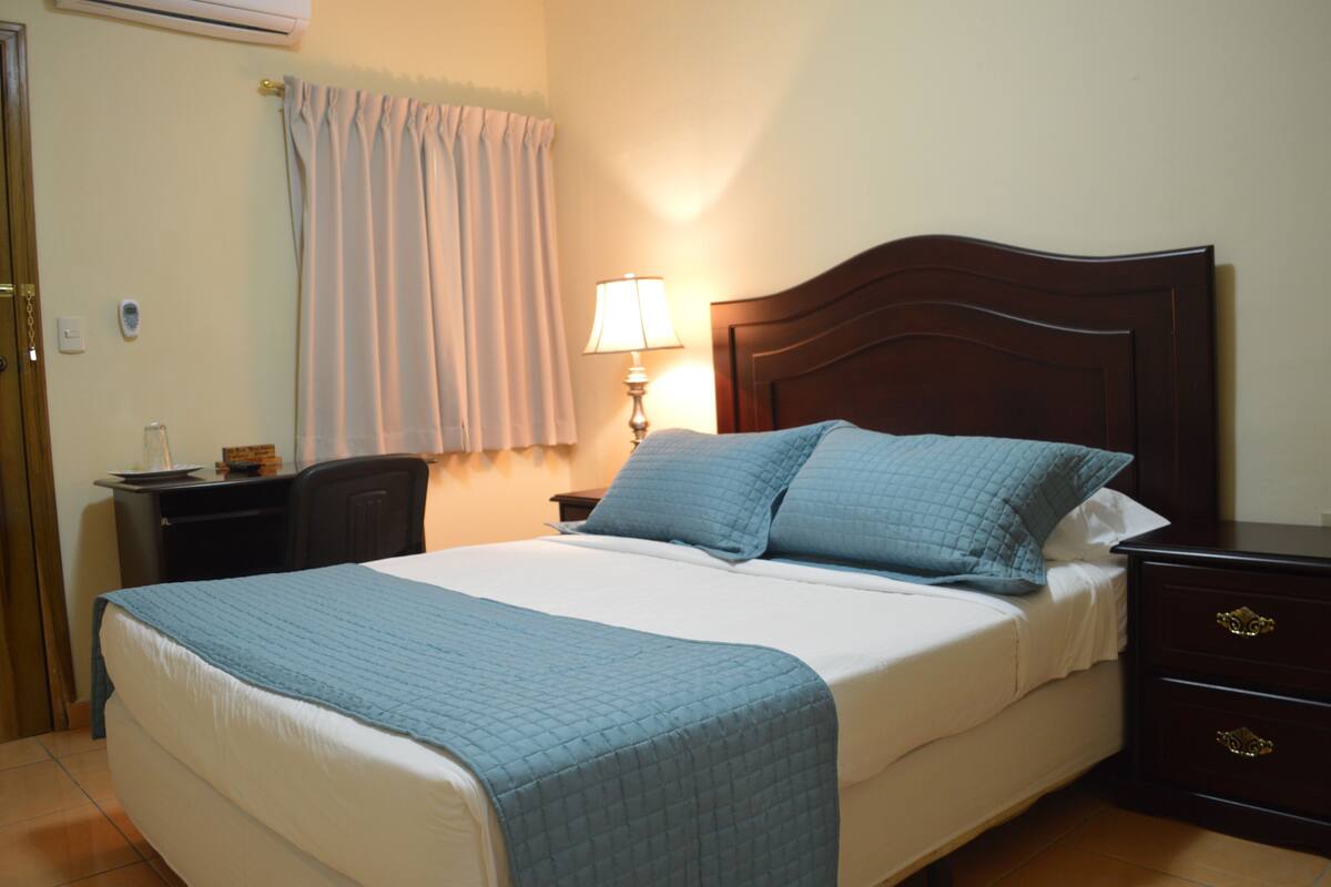 A neatly made bed with a blue quilt, a bedside lamp, and a wooden nightstand with drawers.