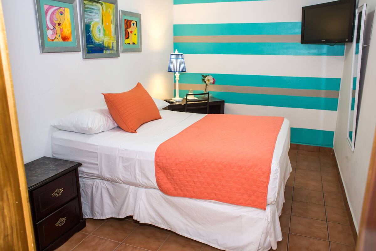 A double bed with white and orange bedding, a nightstand with a lamp, and a wall with striped decor.