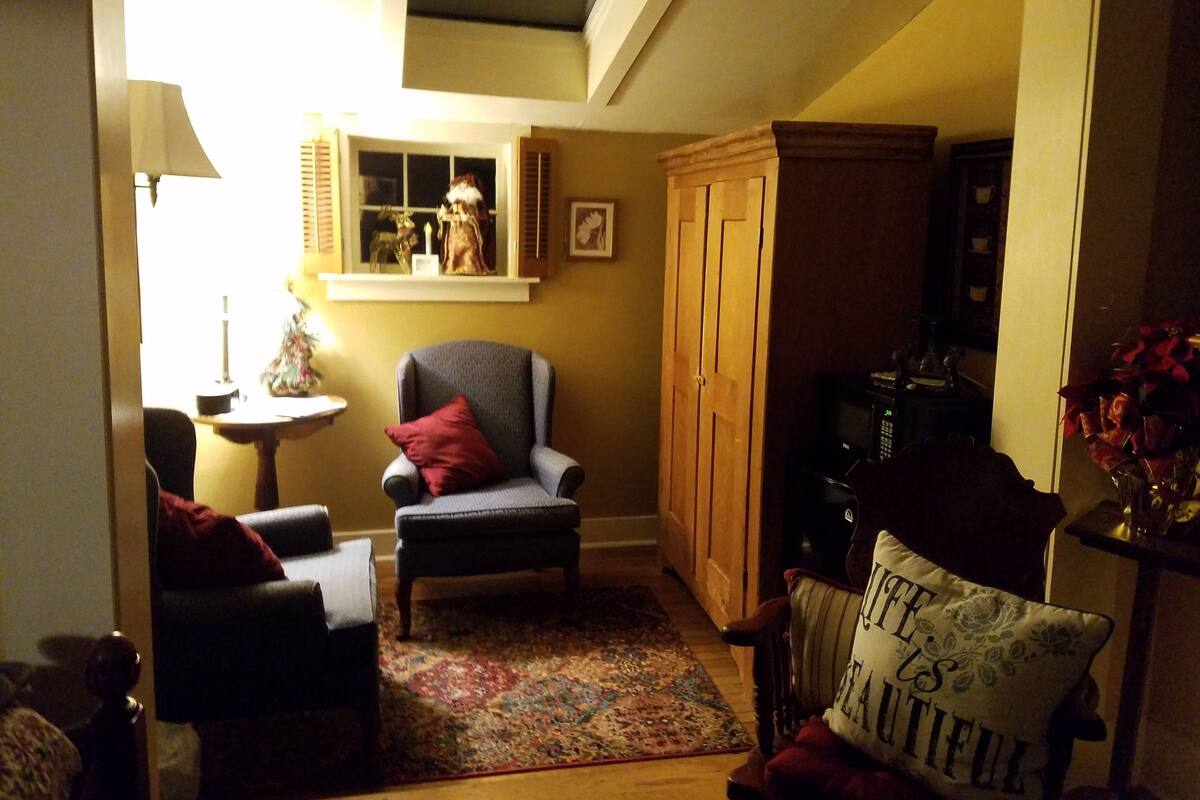 A cozy living room with a high ceiling, a wooden cabinet, a small table with a lamp, and a chair with a red pillow.