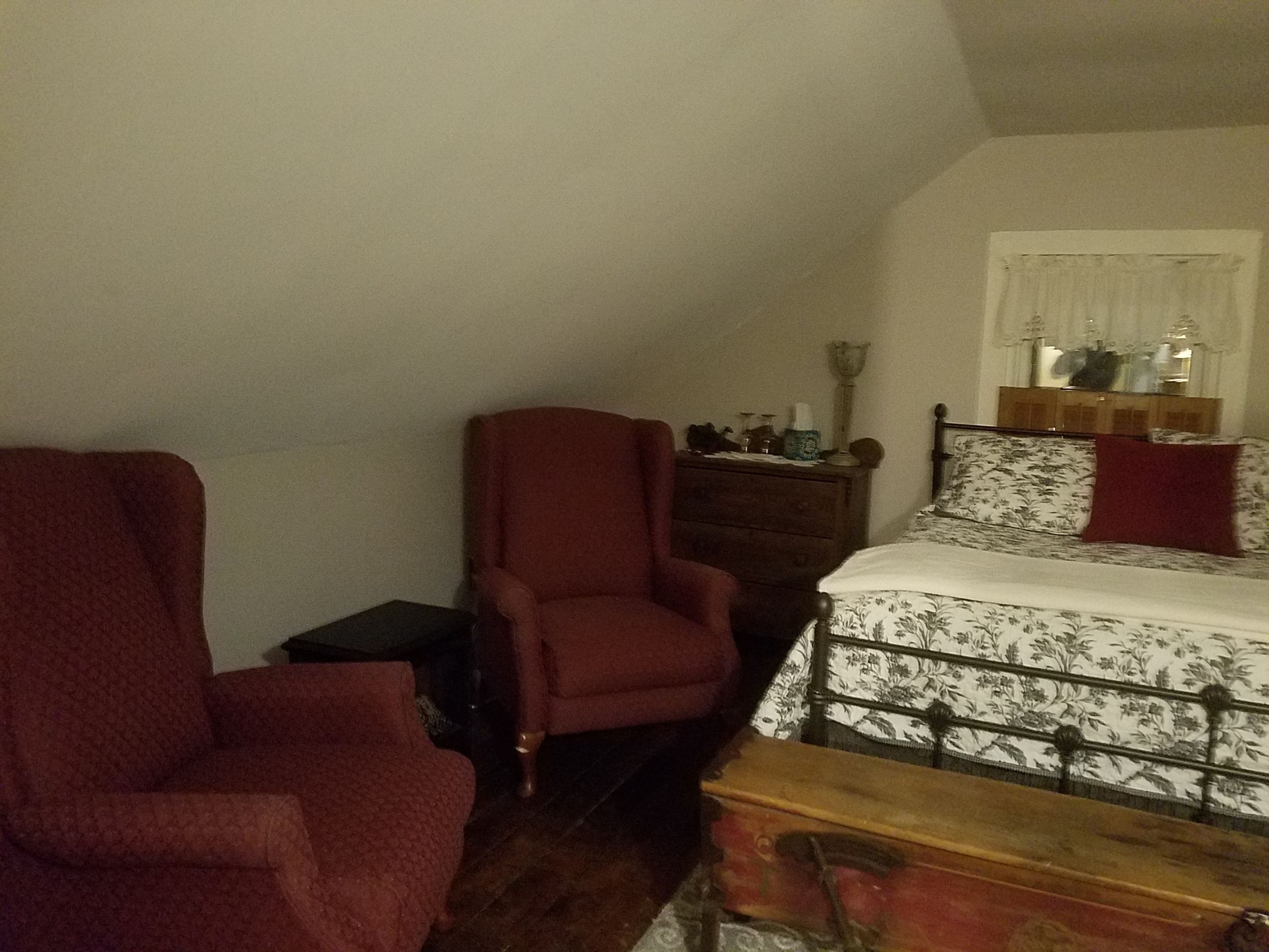 A room with a bed, a red armchair, a wooden chest, and a nightstand with a lamp.