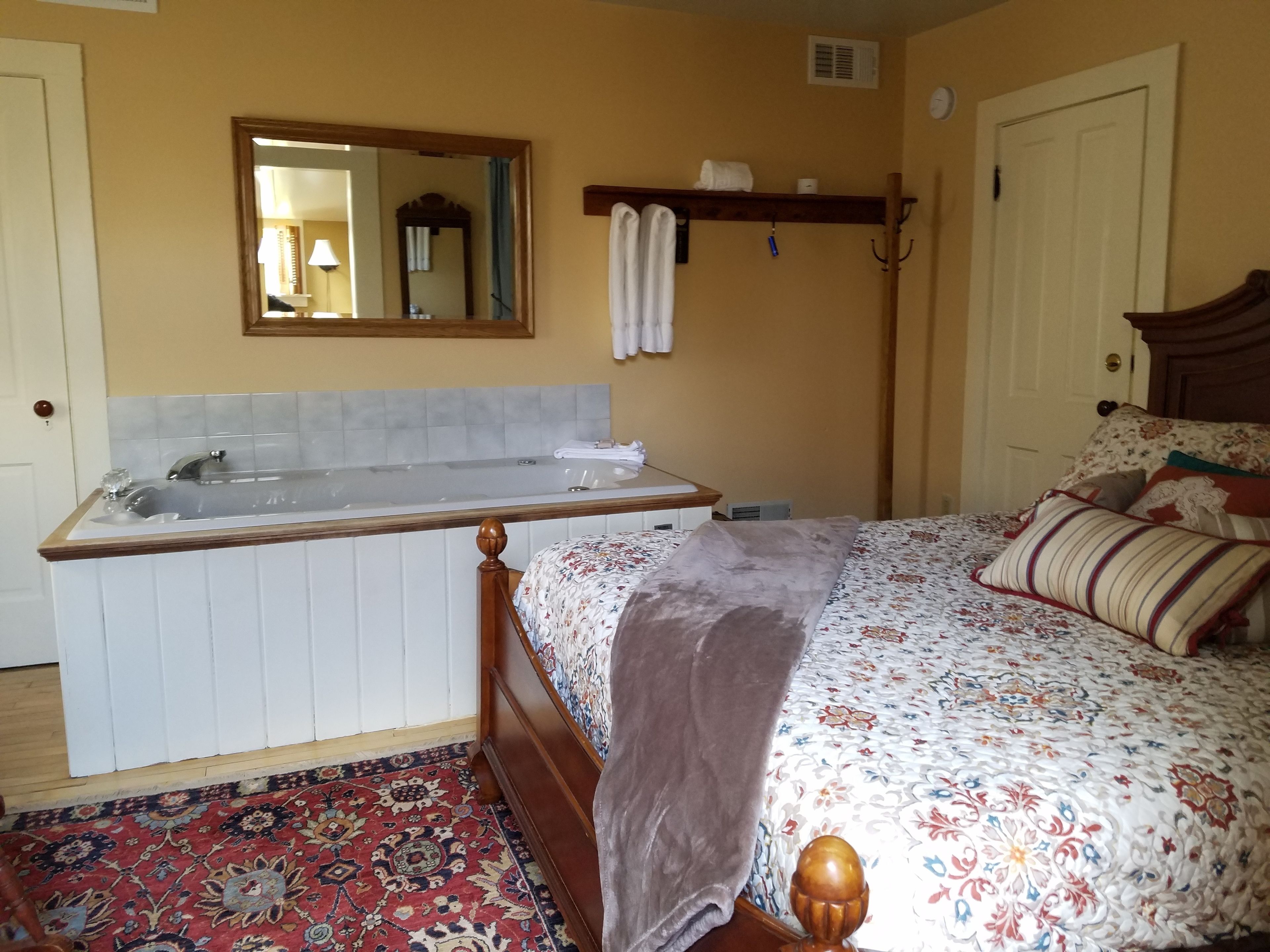 Une chambre avec un grand lit, une tête de lit en bois, une table de chevet, un miroir, une fenêtre et une salle de bain avec une baignoire.