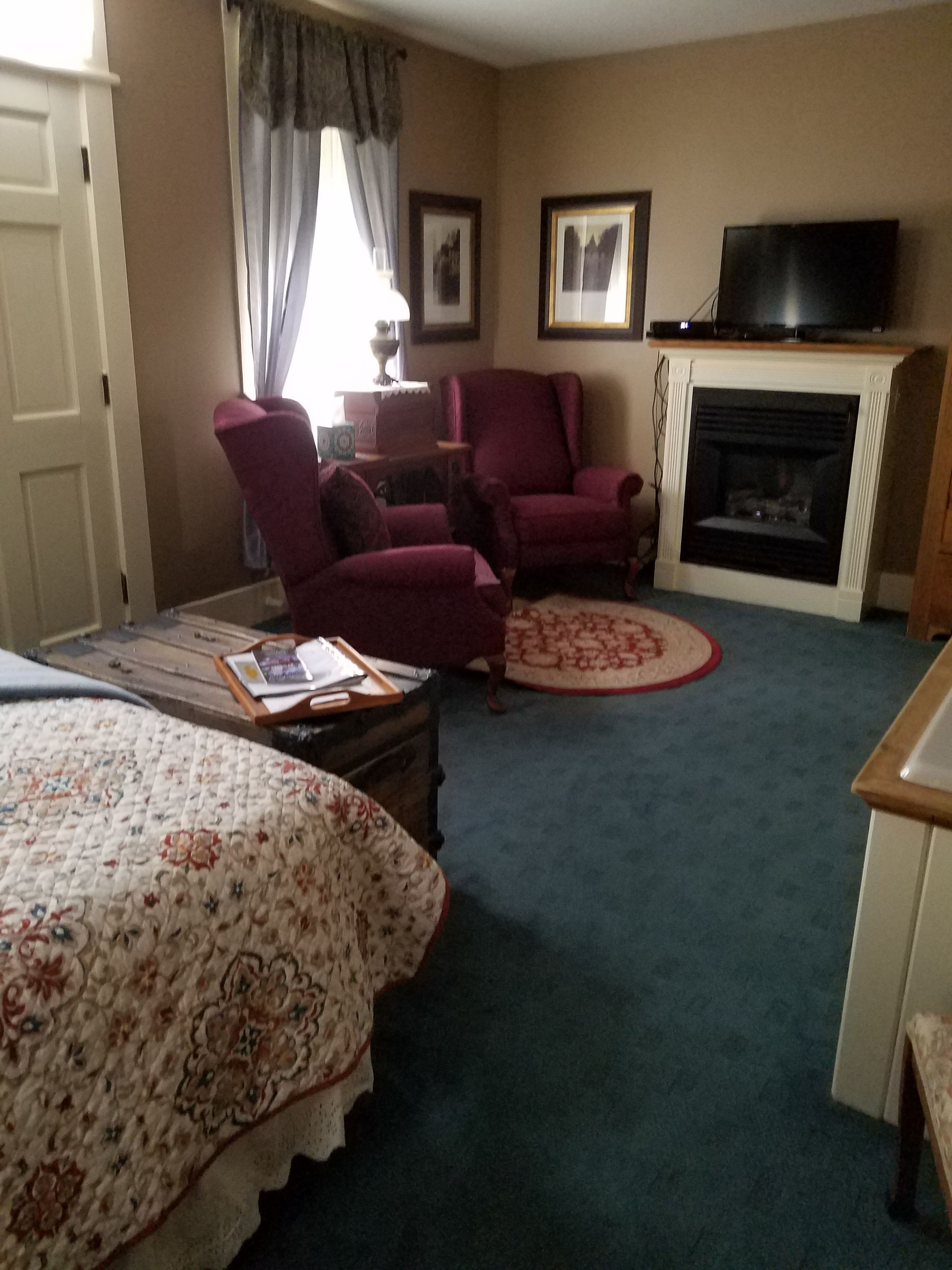 A hotel room with a bed, two armchairs, a small table with a book, a fireplace, and a television on a stand.