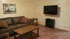 A cozy living room with a brown leather sofa, two matching cushions, a wooden coffee table, a small wooden side table, a black TV mounted on the wall, and a black wood-burning stove.