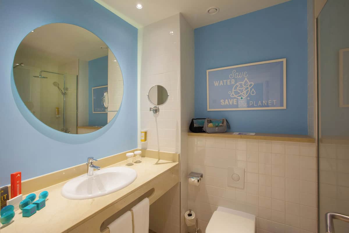 Une salle de bain avec un miroir rond, un lavabo, une douche et un porte-serviettes. Il y a une affiche sur le mur qui dit : « Économisez l’eau, protégez la planète ».