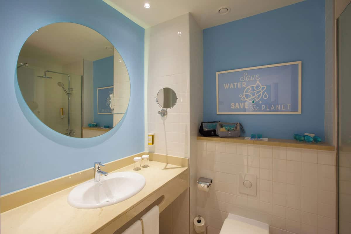 Une salle de bain avec un miroir rond, un lavabo et un porte-serviettes fixé au mur. Un panneau encadré sur le mur porte l’inscription : « Économisez l’eau, préservez la planète. »