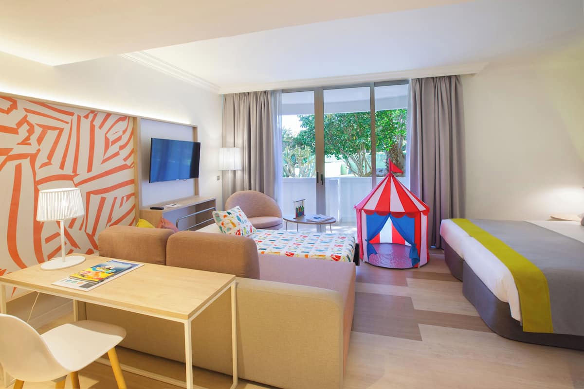 Une chambre d’hôtel comprenant un lit, un bureau, une chaise, un canapé, une télévision, une fenêtre avec des rideaux et une tente de jeu aux couleurs vives.