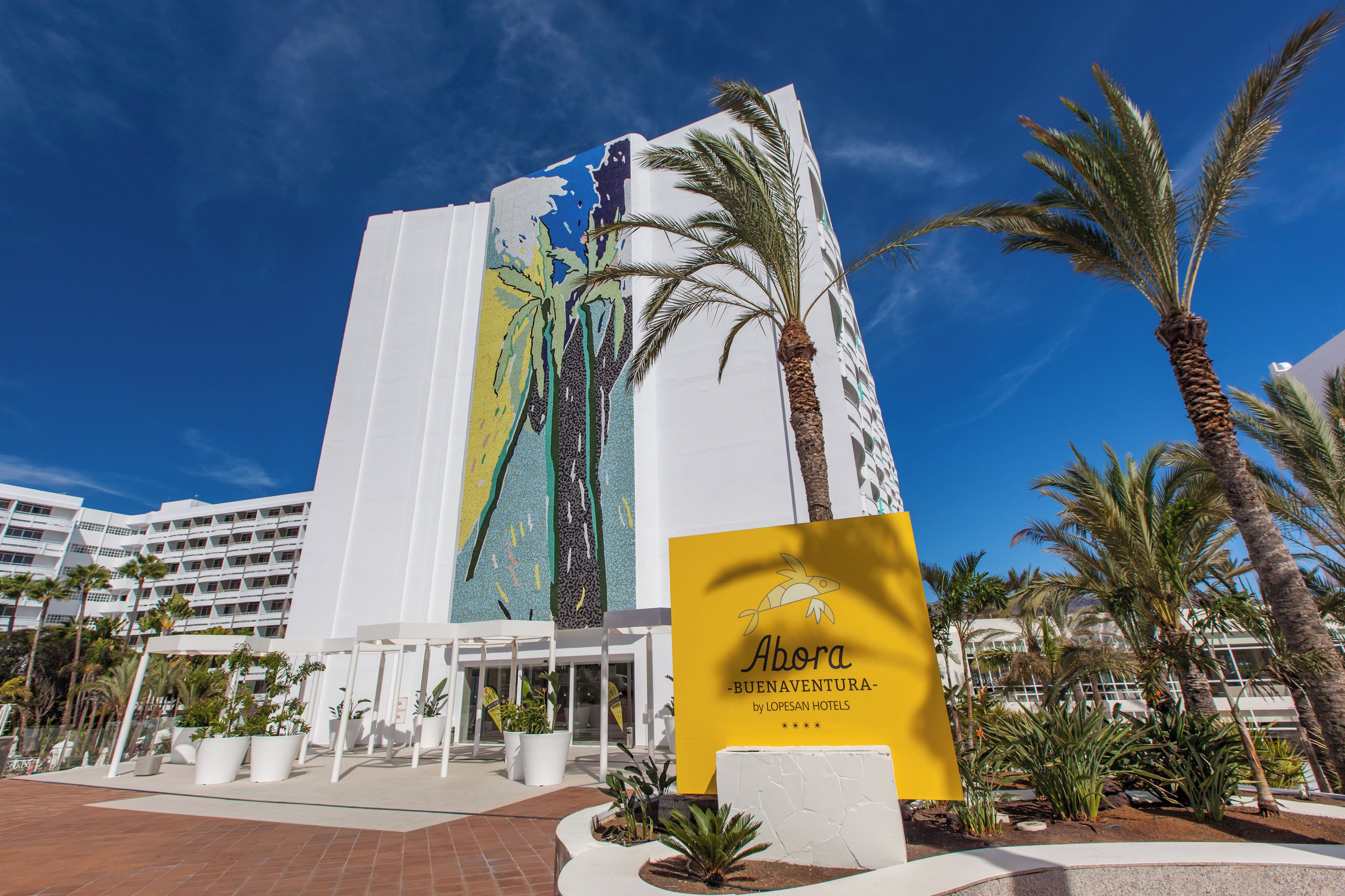 Un hotel con un gran mural en la fachada, palmeras y un cartel que dice 'Abora Buenaventura by LOPESAN HOTELS'.