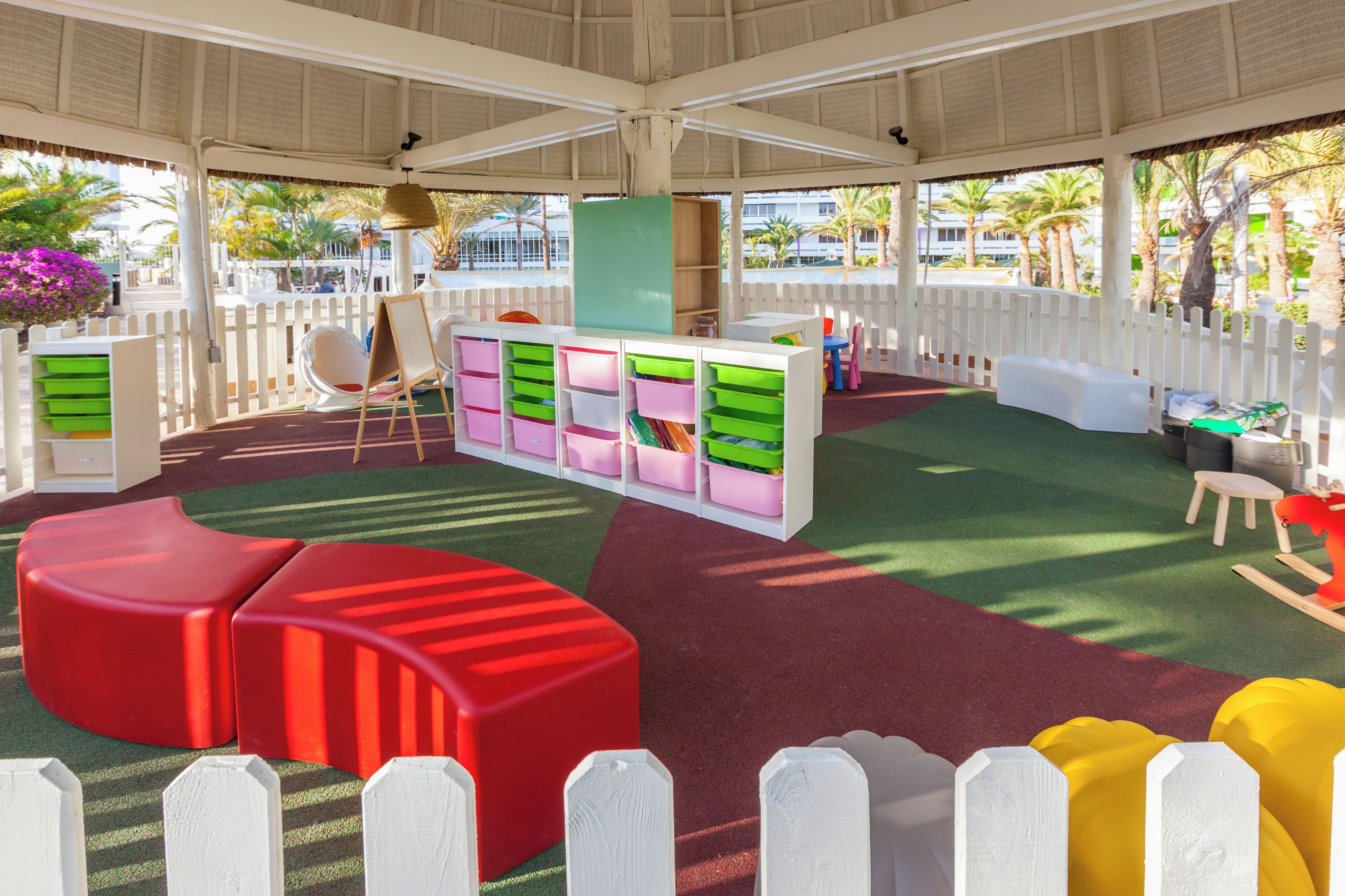 An indoor play area with colorful storage bins, a red play structure, and a white fence.