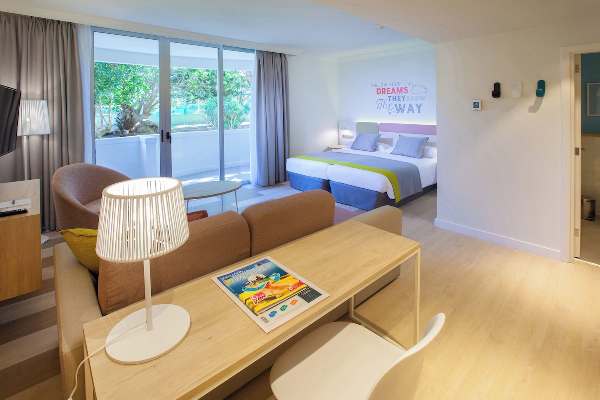 Une chambre d’hôtel moderne avec une grande fenêtre, un lit doté d’une tête de lit colorée, un canapé, une petite table avec une lampe et un mur orné d’un texte motivant.