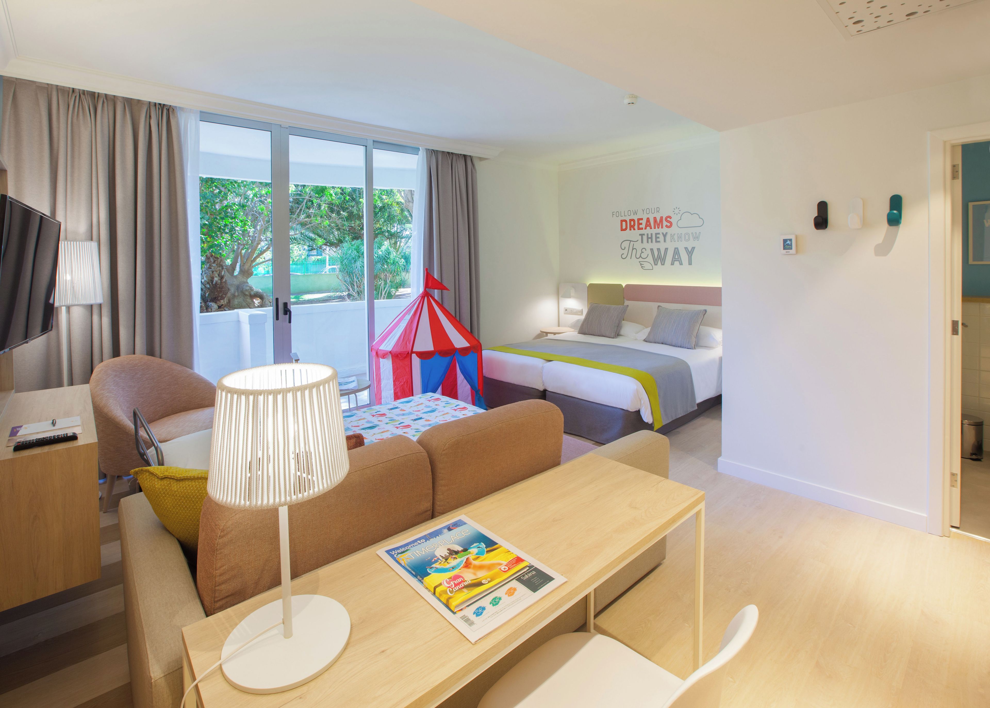 Habitación de hotel con cama, escritorio, silla, lámpara, ventana con cortinas y una pared con una frase motivacional.
