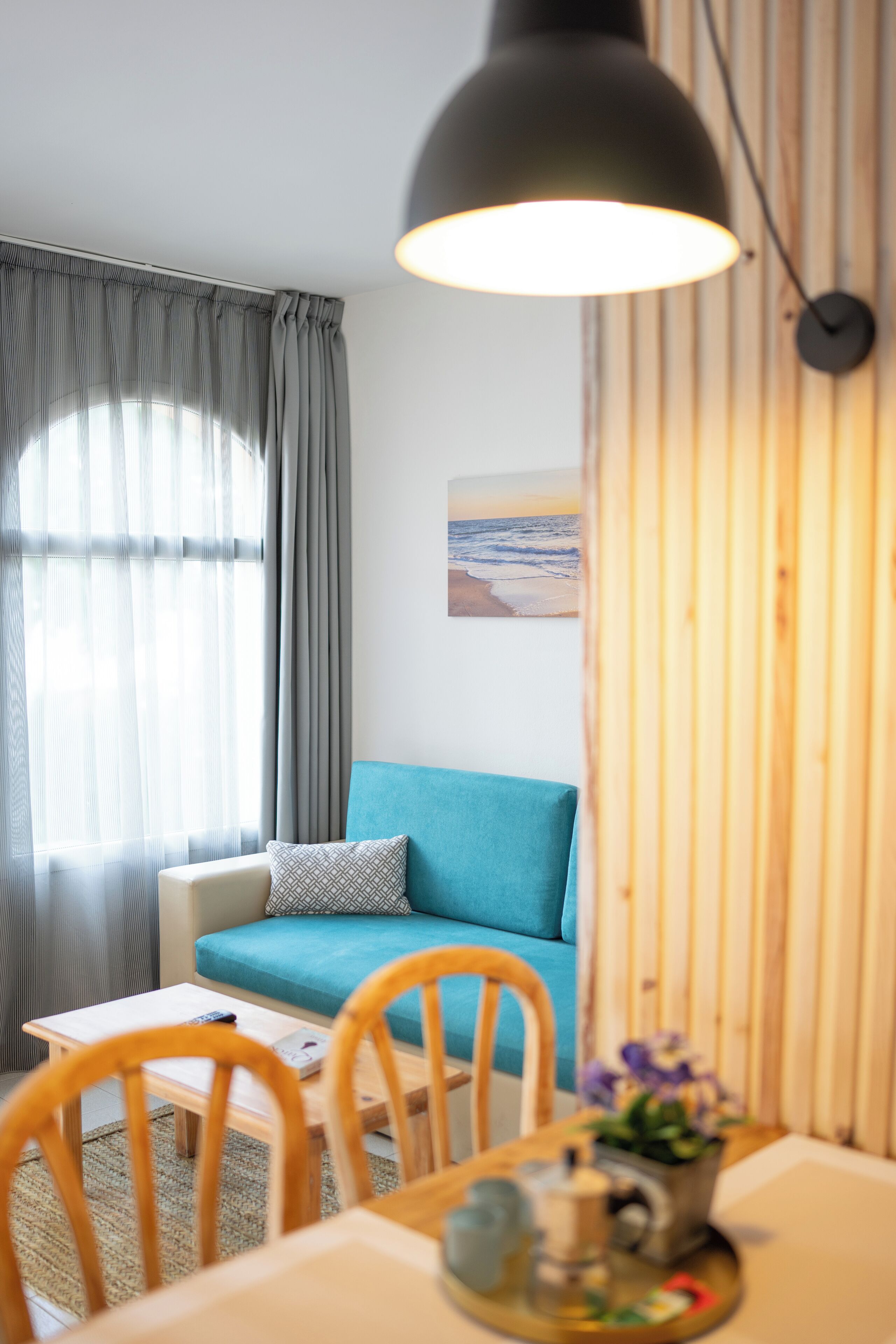 A cozy living room with a teal sofa, a wooden dining table with chairs, a coffee table, and a pendant light.