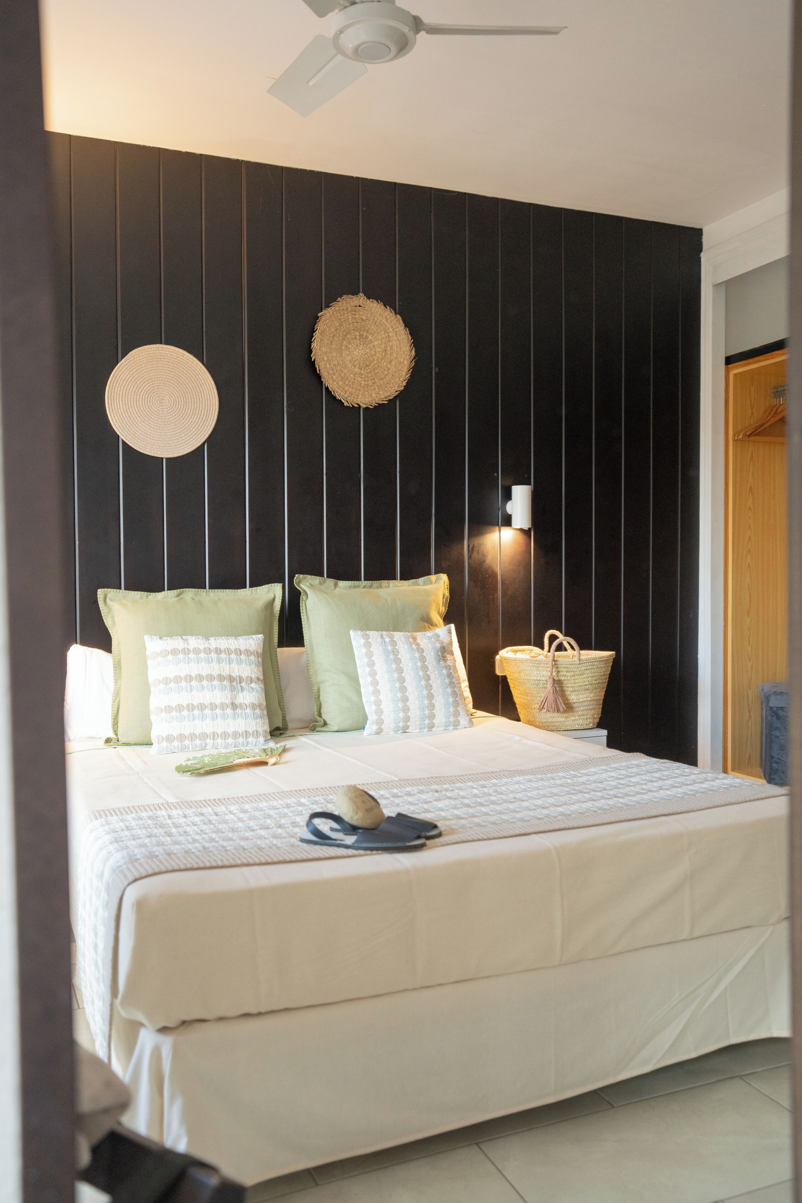 A neatly made bed with pillows, a woven wall hanging, and a basket on a dark wooden wall.