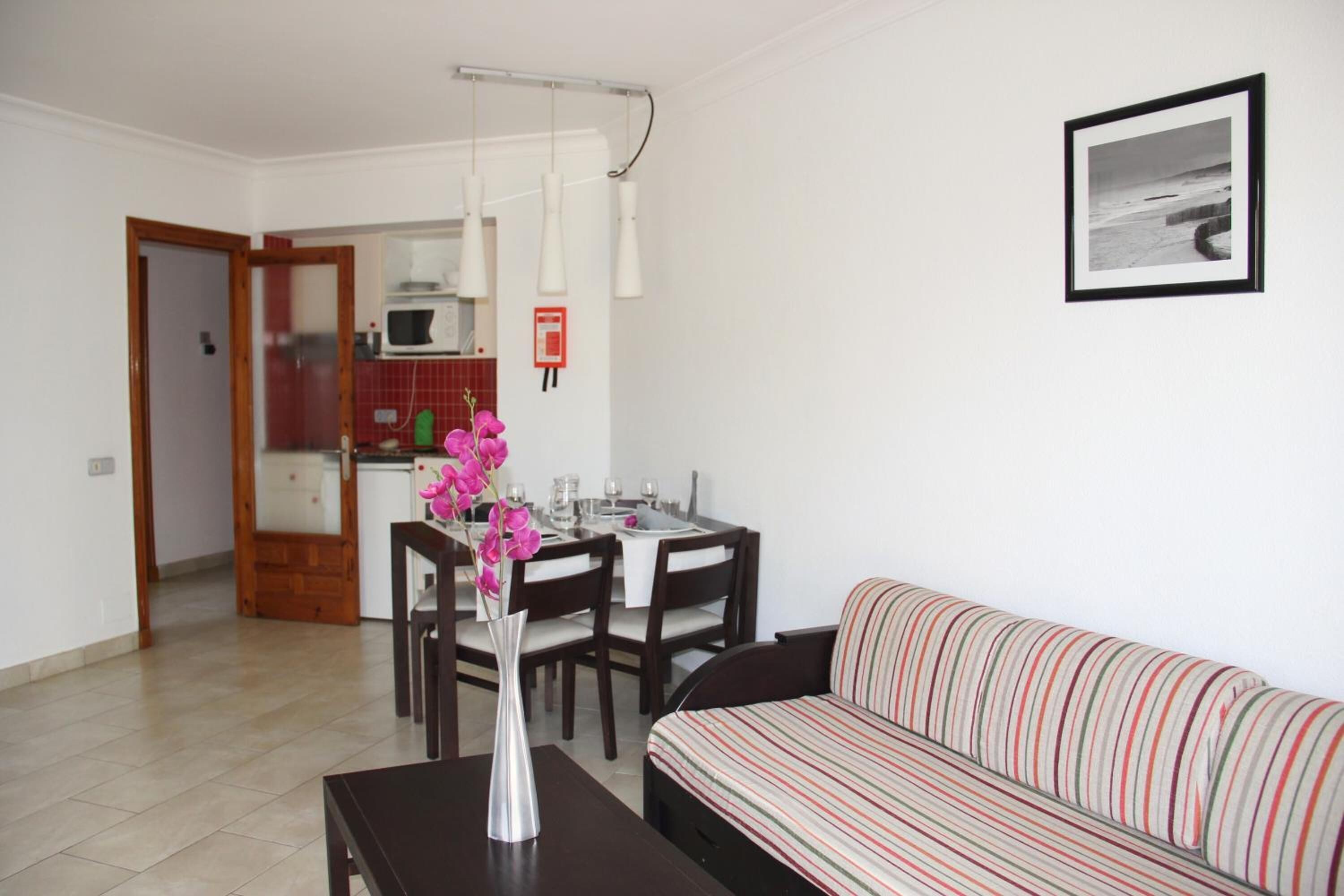 Una sala de estar con un sofá a rayas, una mesa de comedor con sillas, un florero con flores y una cocina al fondo.