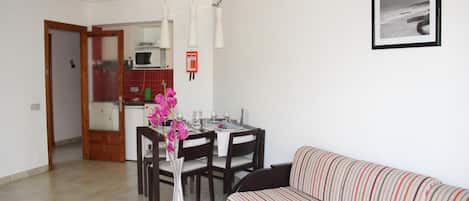 A living room with a striped sofa, a dining table with chairs, a vase with flowers, and a kitchen in the background.