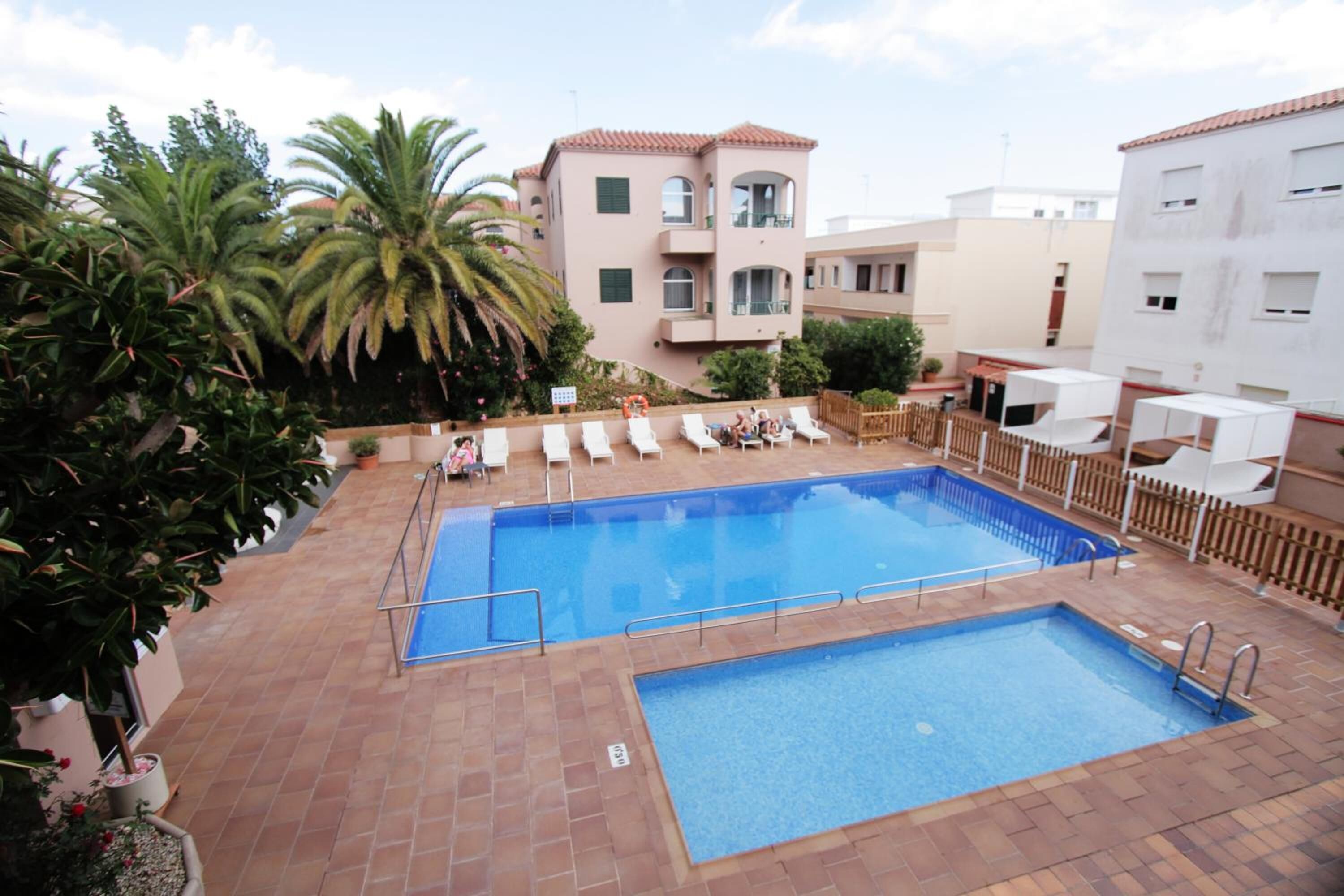 A rooftop swimming pool with lounge chairs, palm trees, and a building with a red-tiled roof.