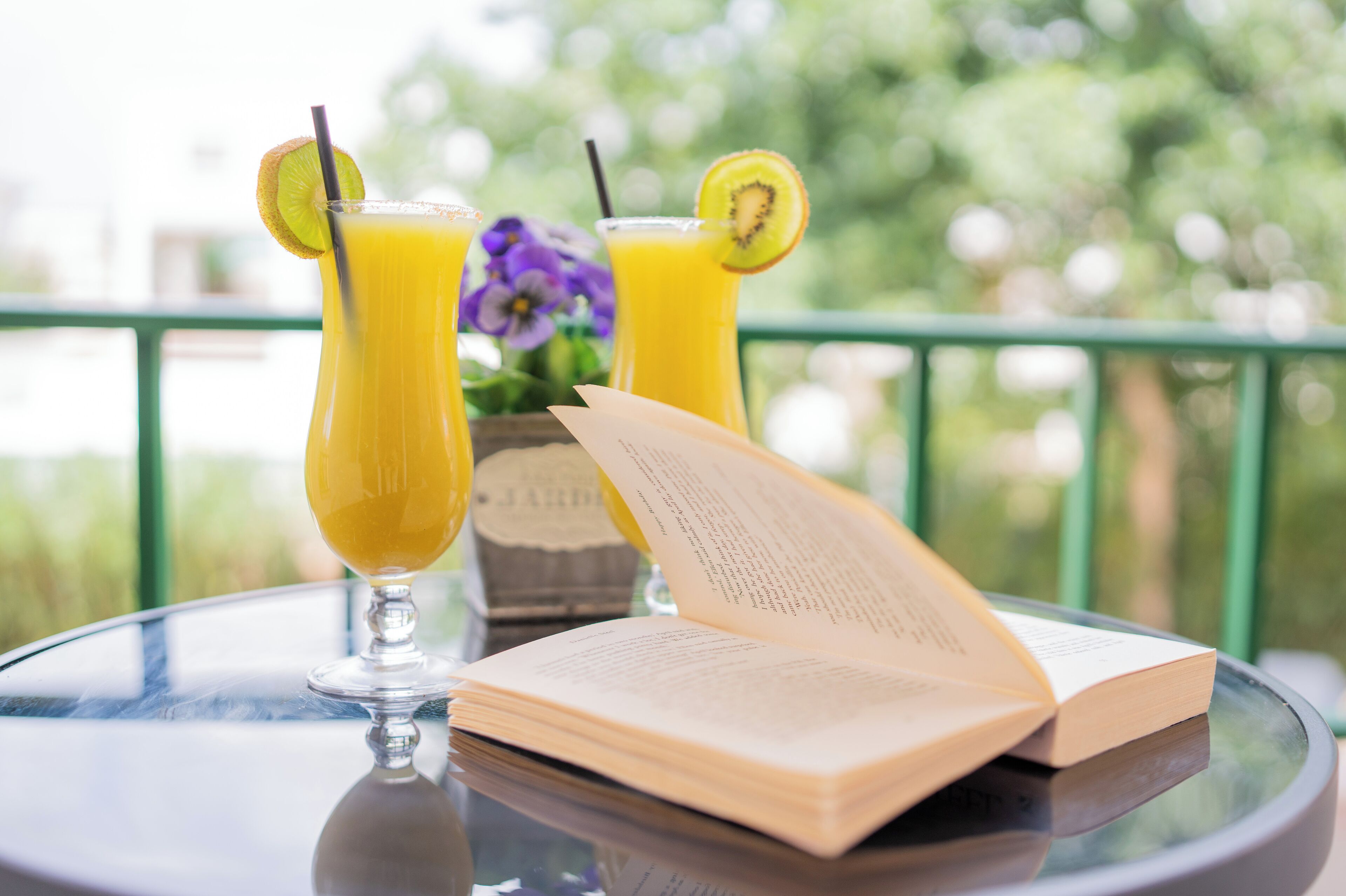 Dos vasos de jugo de naranja con limón y rodajas de kiwi, un libro y un florero con flores sobre una mesa de vidrio.