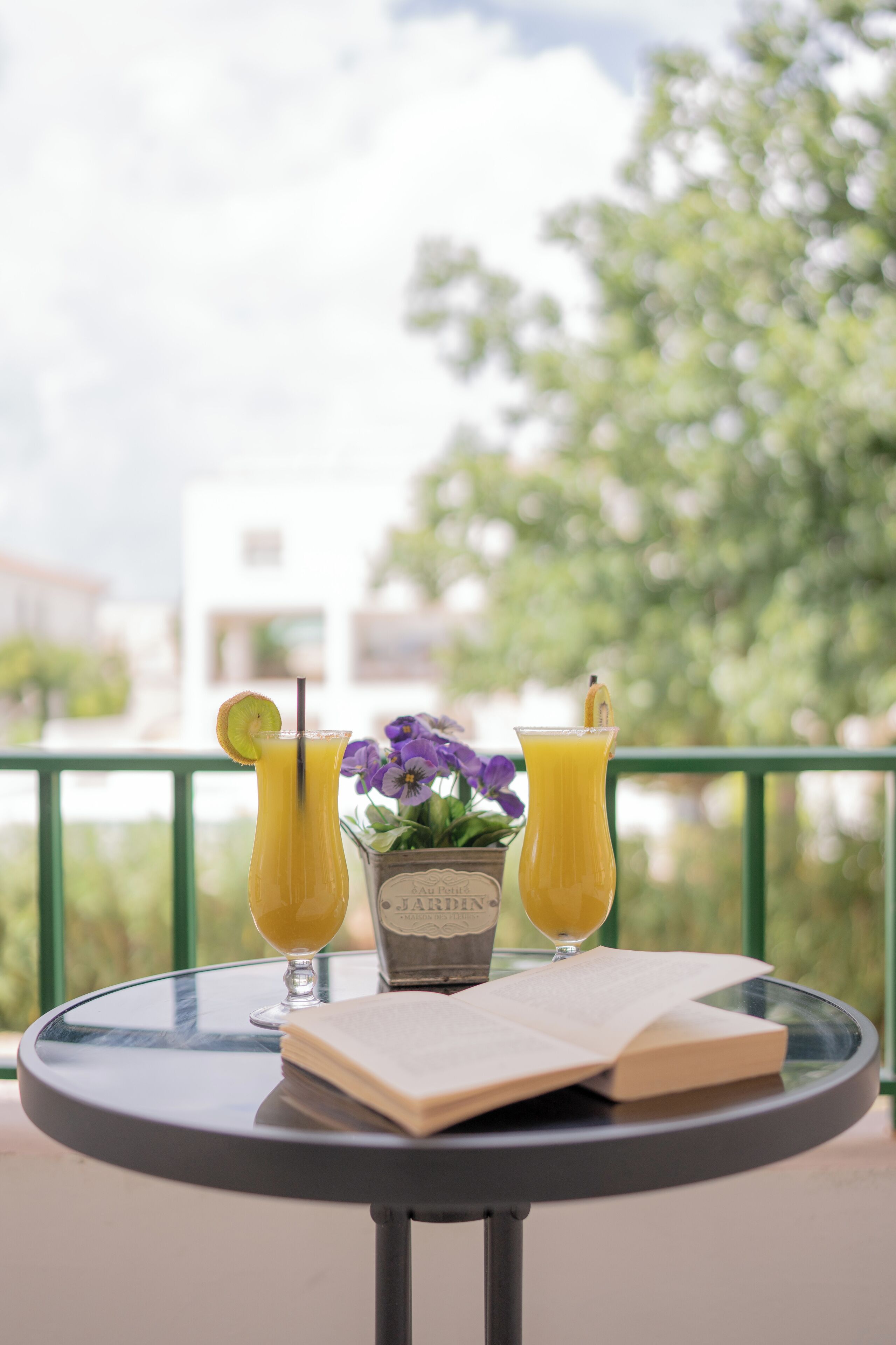Una mesa con dos bebidas, un libro y un florero en un balcón con vistas a árboles y edificios.