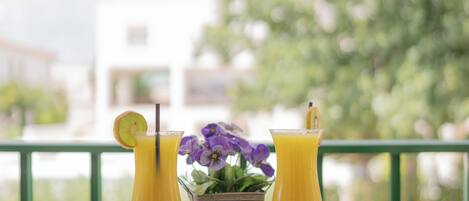 A table with two drinks, a book, and a flower vase on a balcony with a view of trees and buildings.