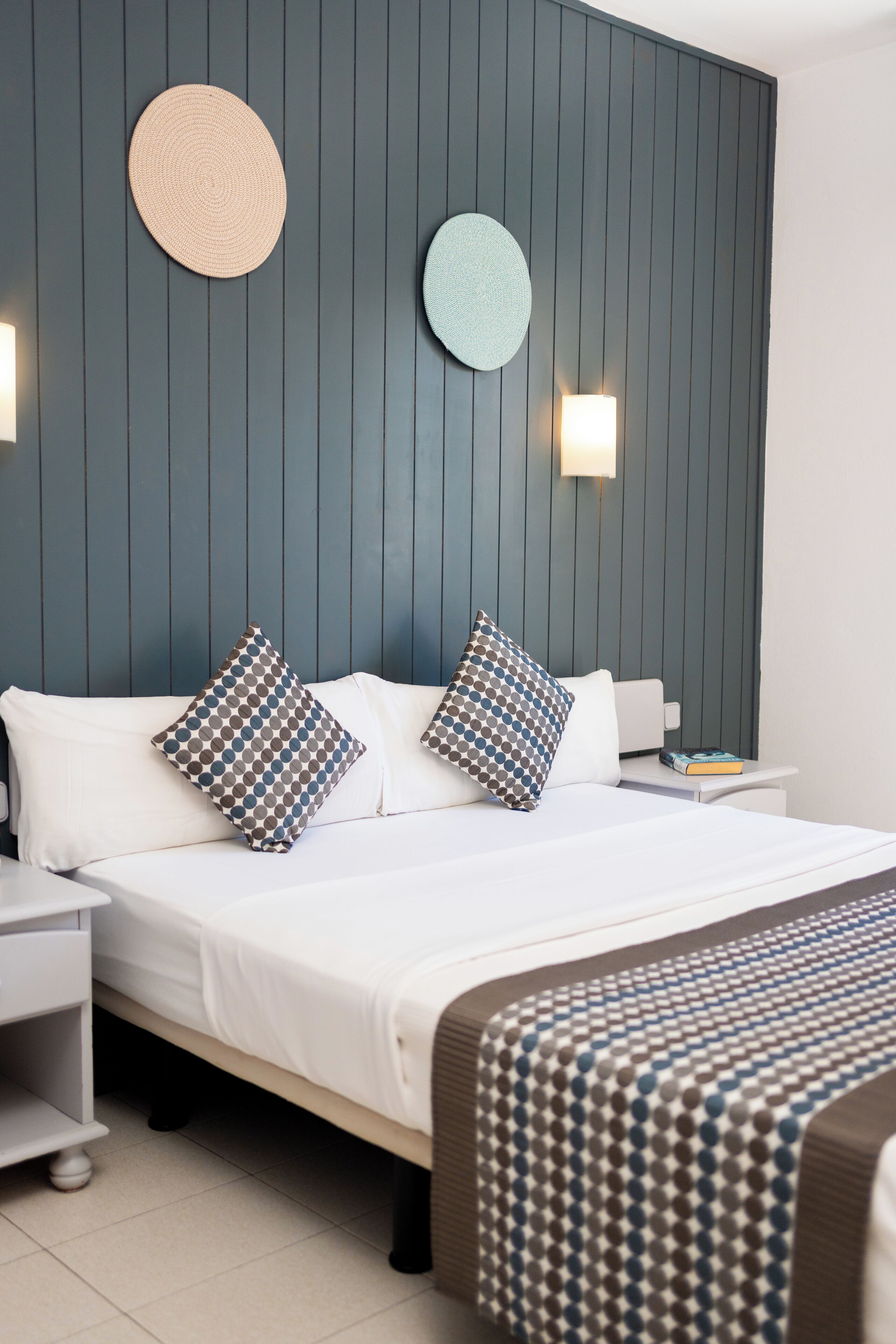 A modern hotel room with a bed, two bedside tables, a nightstand, and a wall-mounted round mirror.