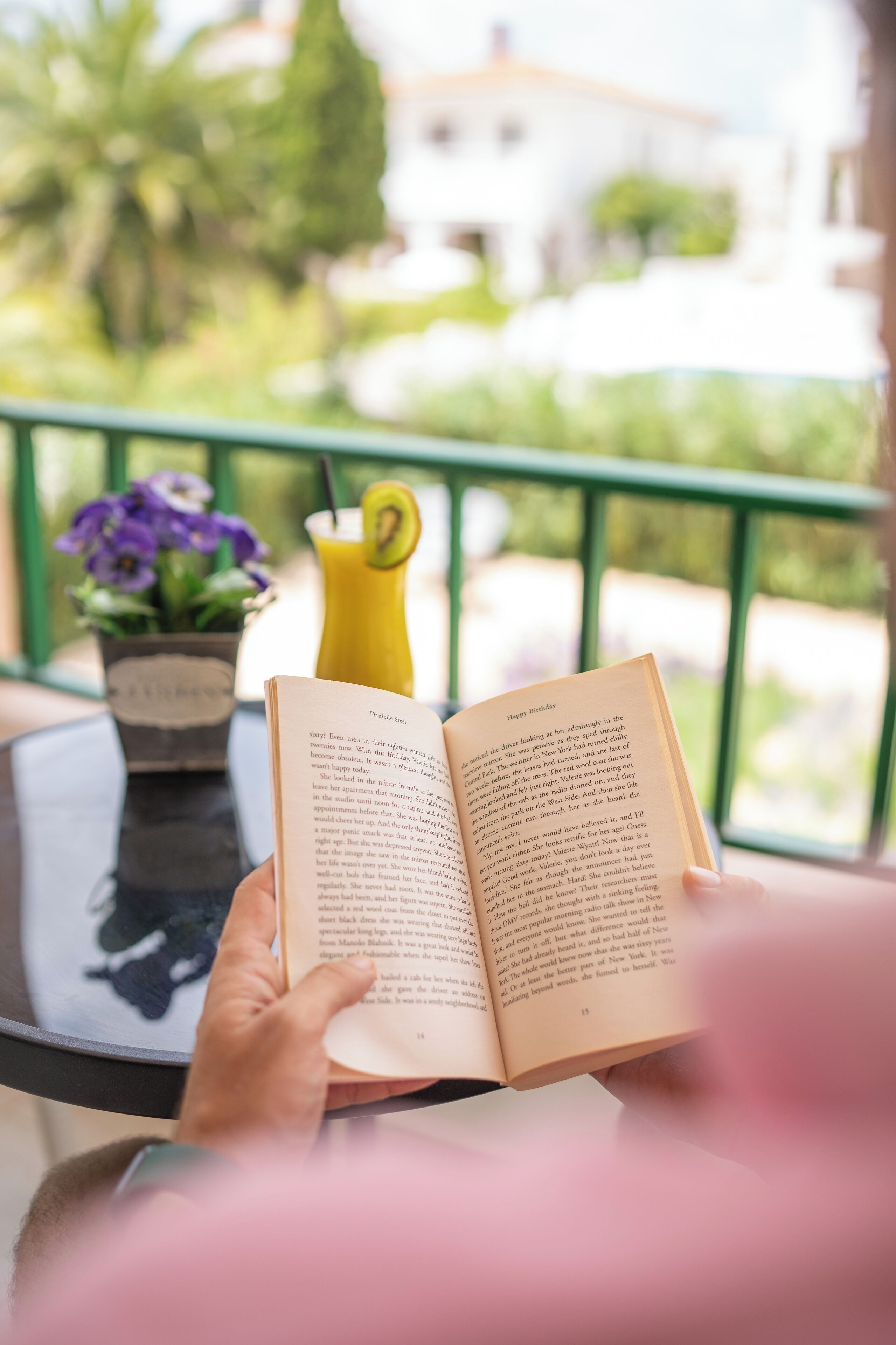 A person reading a book on a balcony with a potted plant and a yellow drink container in the background.
