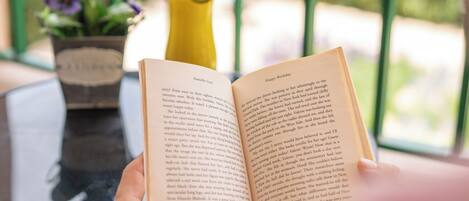 A person reading a book on a balcony with a potted plant and a yellow drink container in the background.