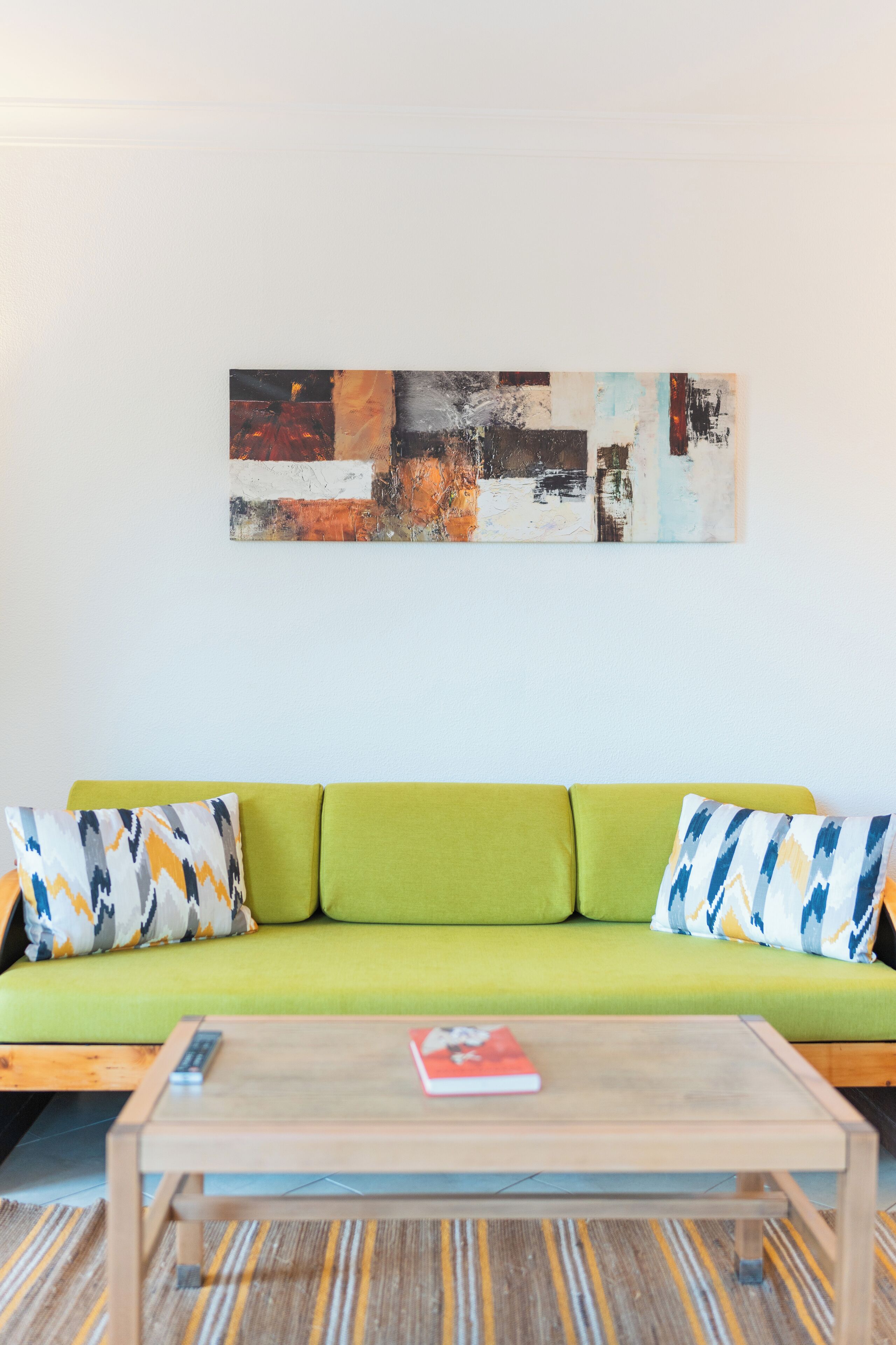 Un sofá verde con cojines estampados, una mesa de centro de madera con un libro, y un cuadro abstracto grande en la pared.