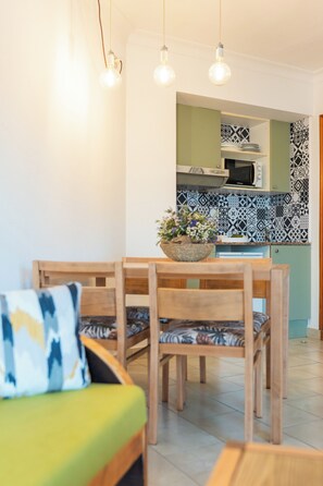 A dining area with a wooden table and chairs, a bowl of flowers, and a green cushion on a sofa. - Apartamentos Royal Life (Mahón)