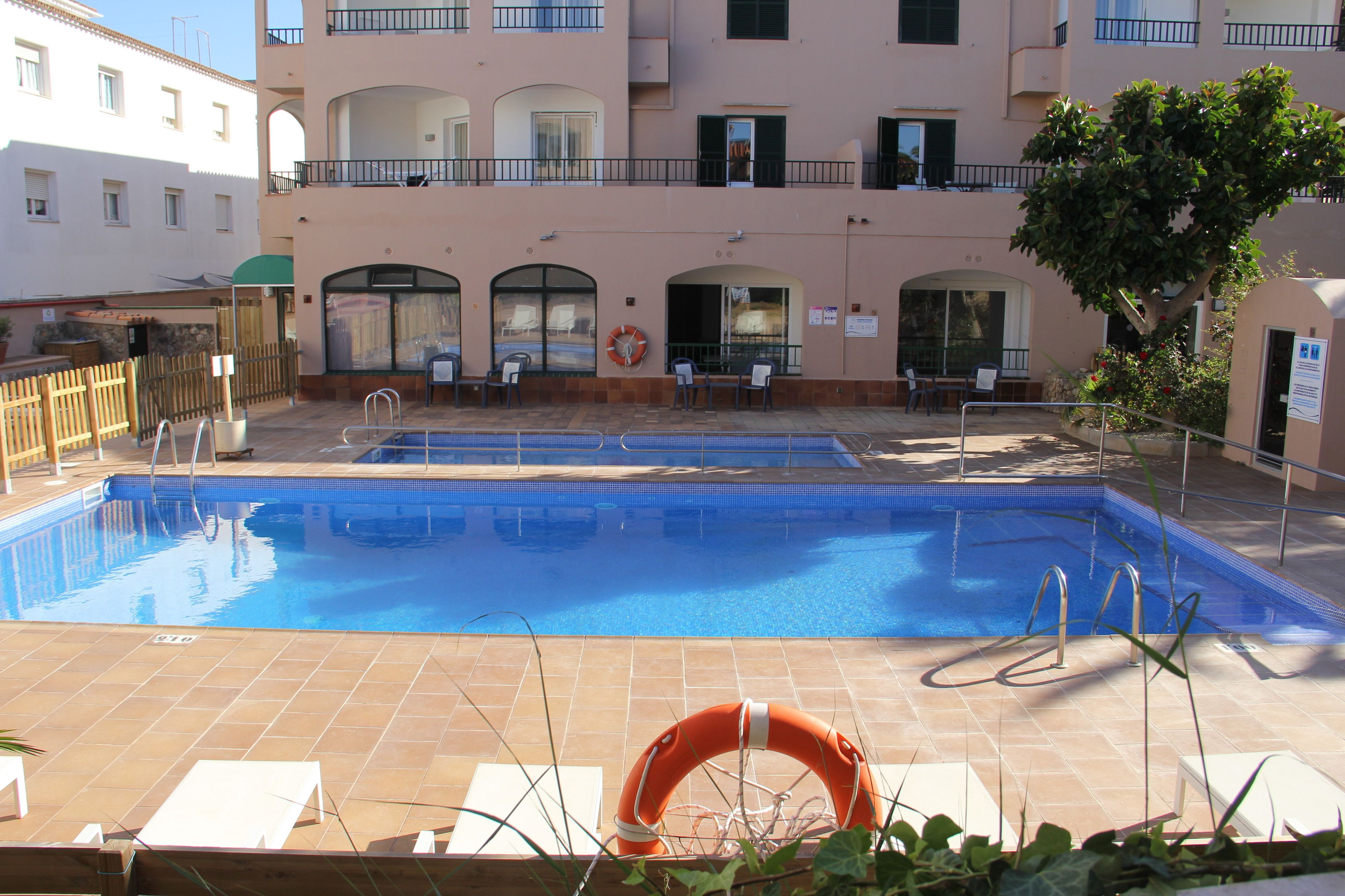 A swimming pool with a lifebuoy, lounge chairs, and a building with balconies in the background.